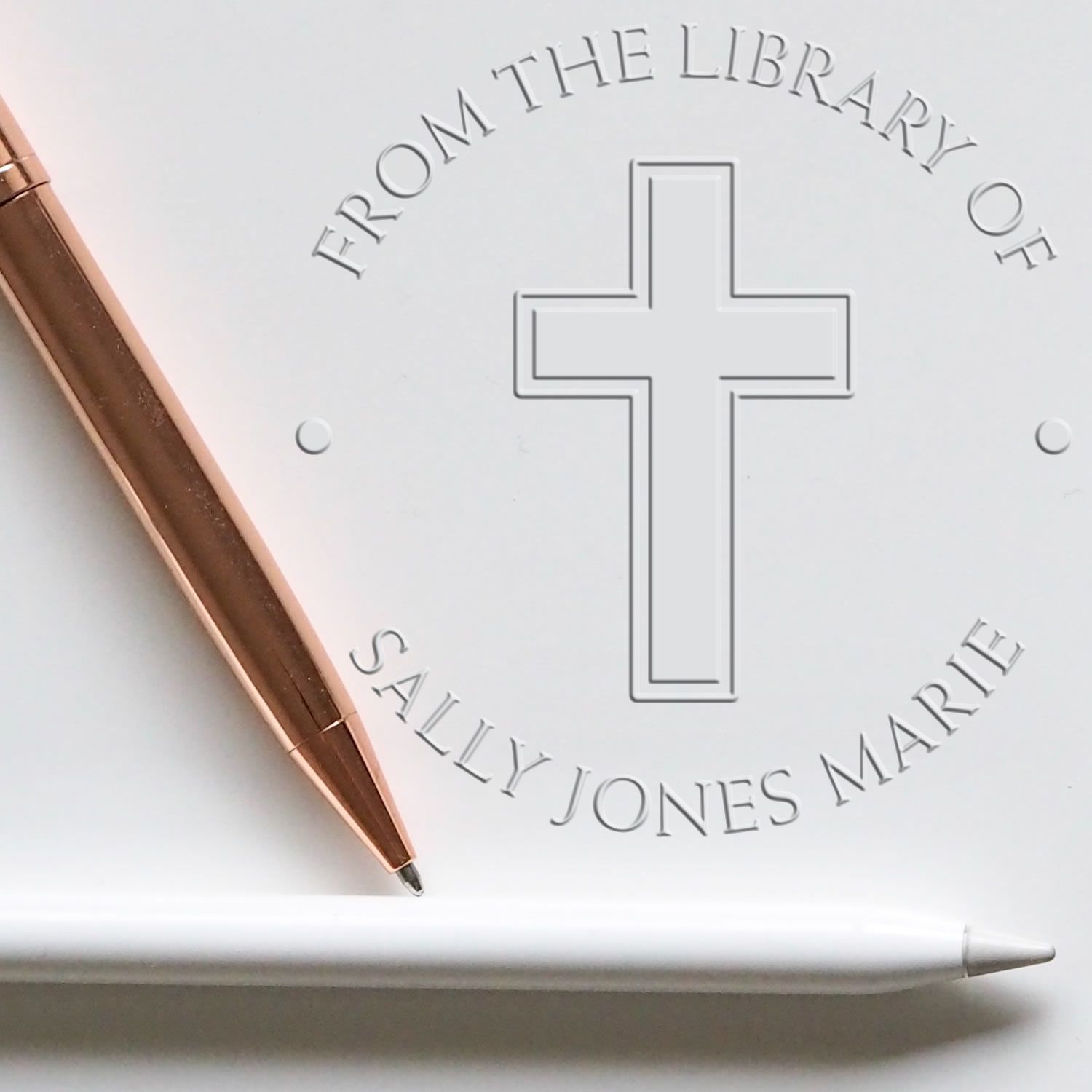 White memorial stamp and seal with silver cross and engraved text, beside a rose gold pen and white stylus.