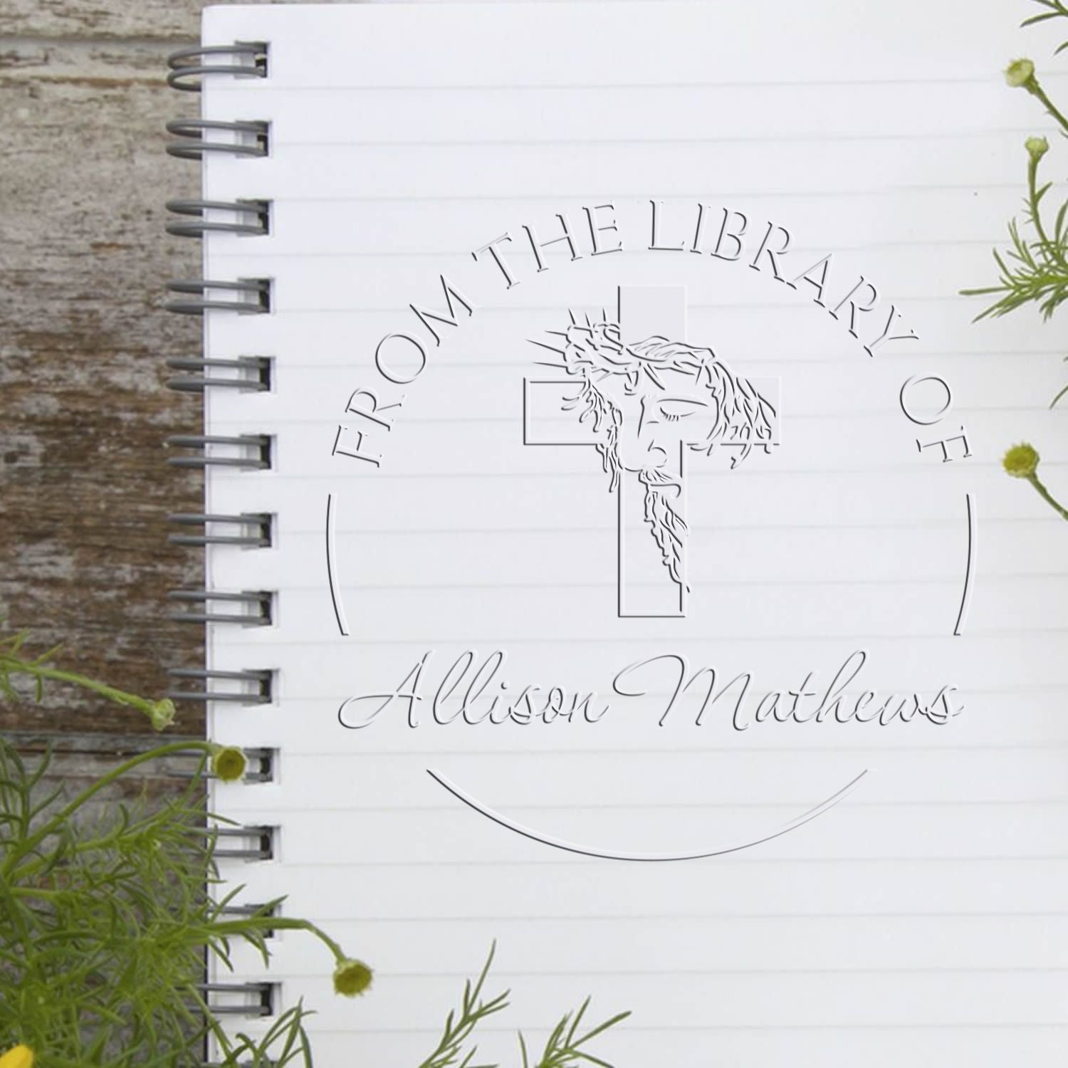 White spiral notebook with a silver custom seal stamp, script logo, and monogram on rustic wood, accented by green sprigs.