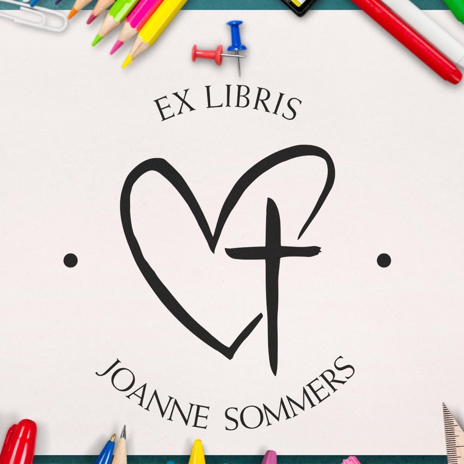 Custom black ex libris heart-and-cross book stamp seal on white paper, framed by colorful pencils and push pins.