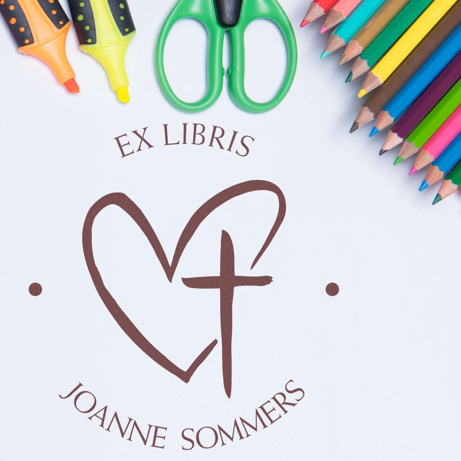 Custom brown ex libris stamp and seal with heart-cross design, surrounded by colorful pencils, scissors, and highlighters on white.