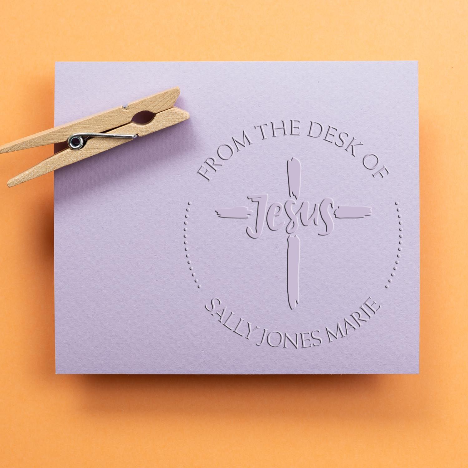 Lavender embossed memorial stamp and seal with cross, Jesus text, and clothespin on a warm orange background.