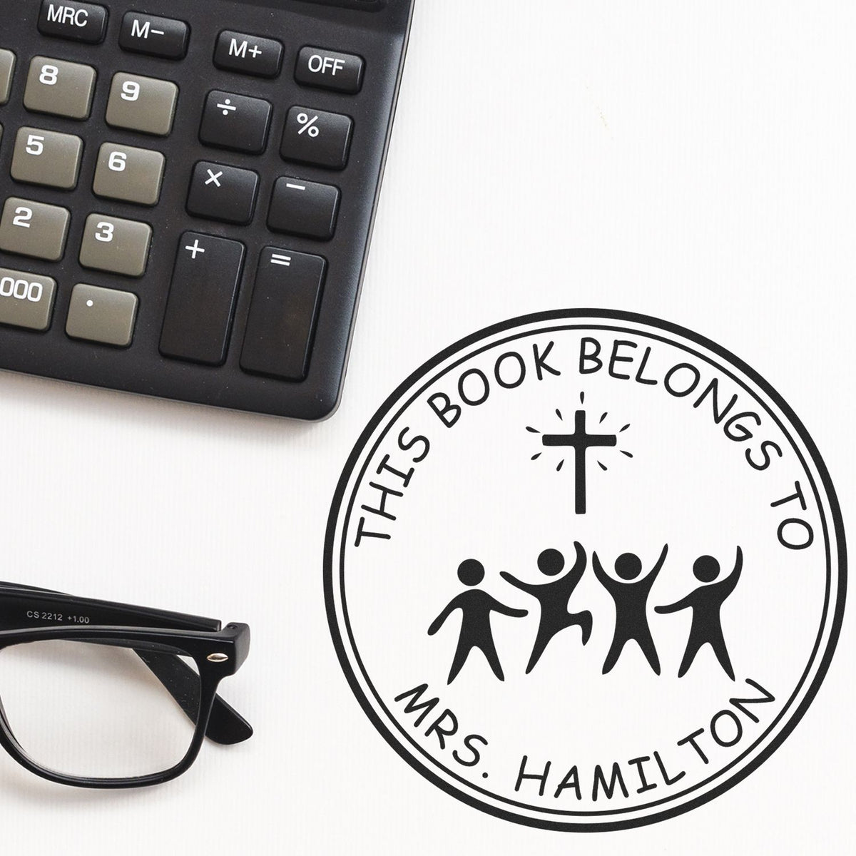 Black custom book-belongings stamp seal with cross and figures, beside a calculator and eyeglasses on white background.