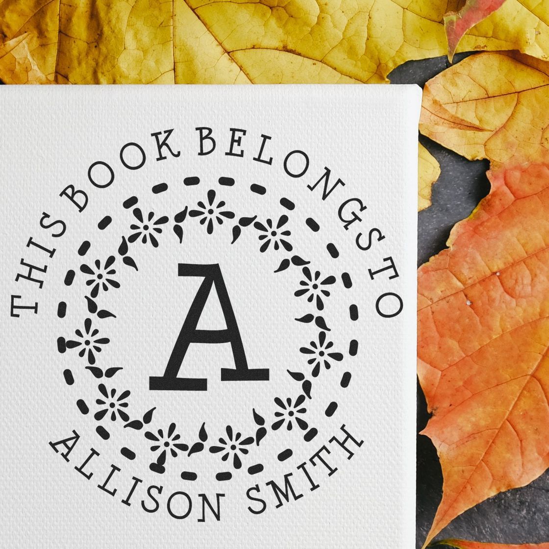 Black custom stamp seal with floral wreath and monogram A on white cardstock, set against yellow and orange autumn leaves.