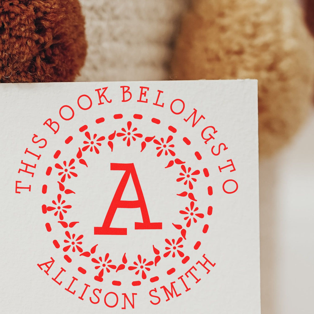 Red personalized book stamp seal with a large A, floral circle, and This Book Belongs to Allison Smith on white paper.