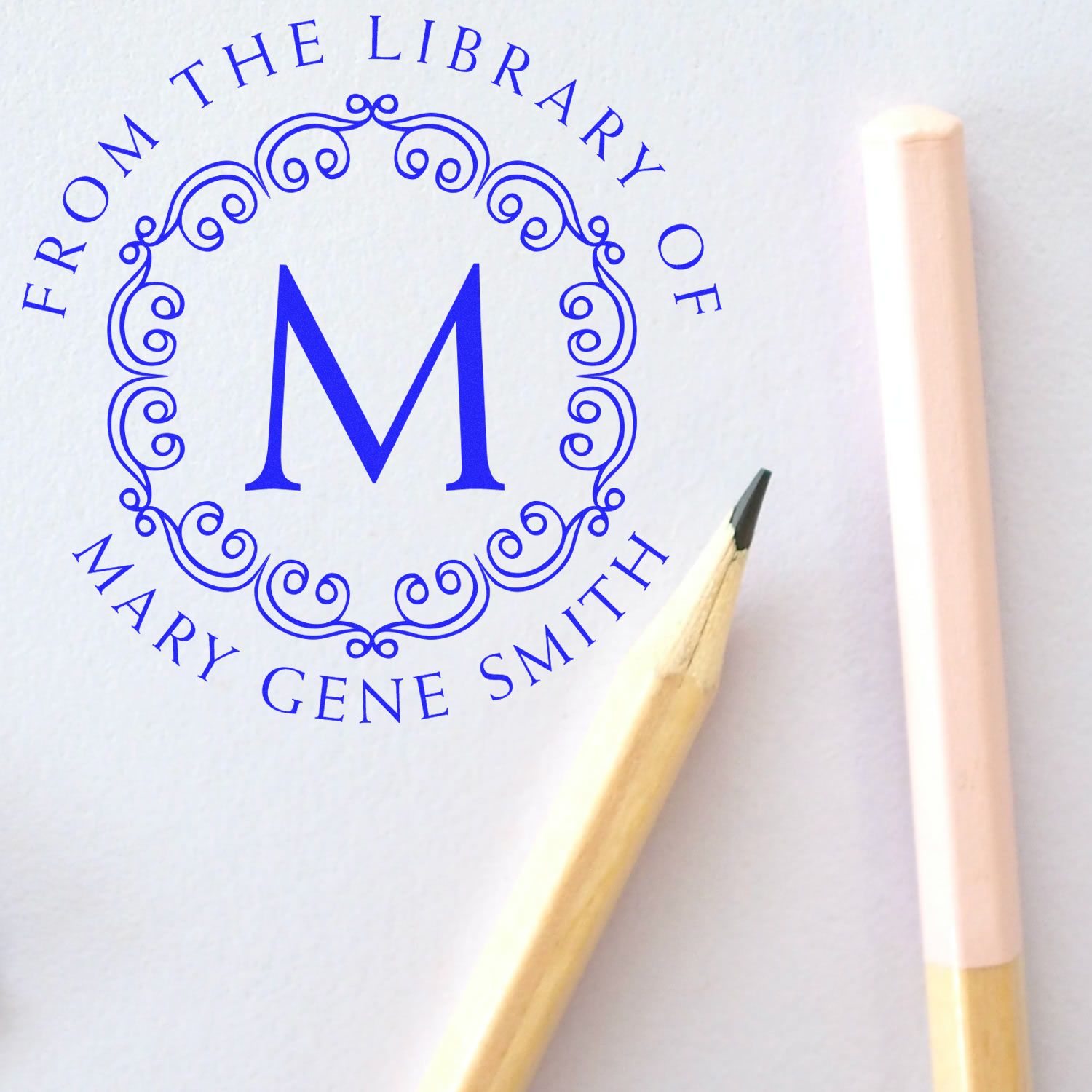 Blue custom library stamp seal with ornate M and From the Library of Mary Gene Smith on white paper, beside a pencil.