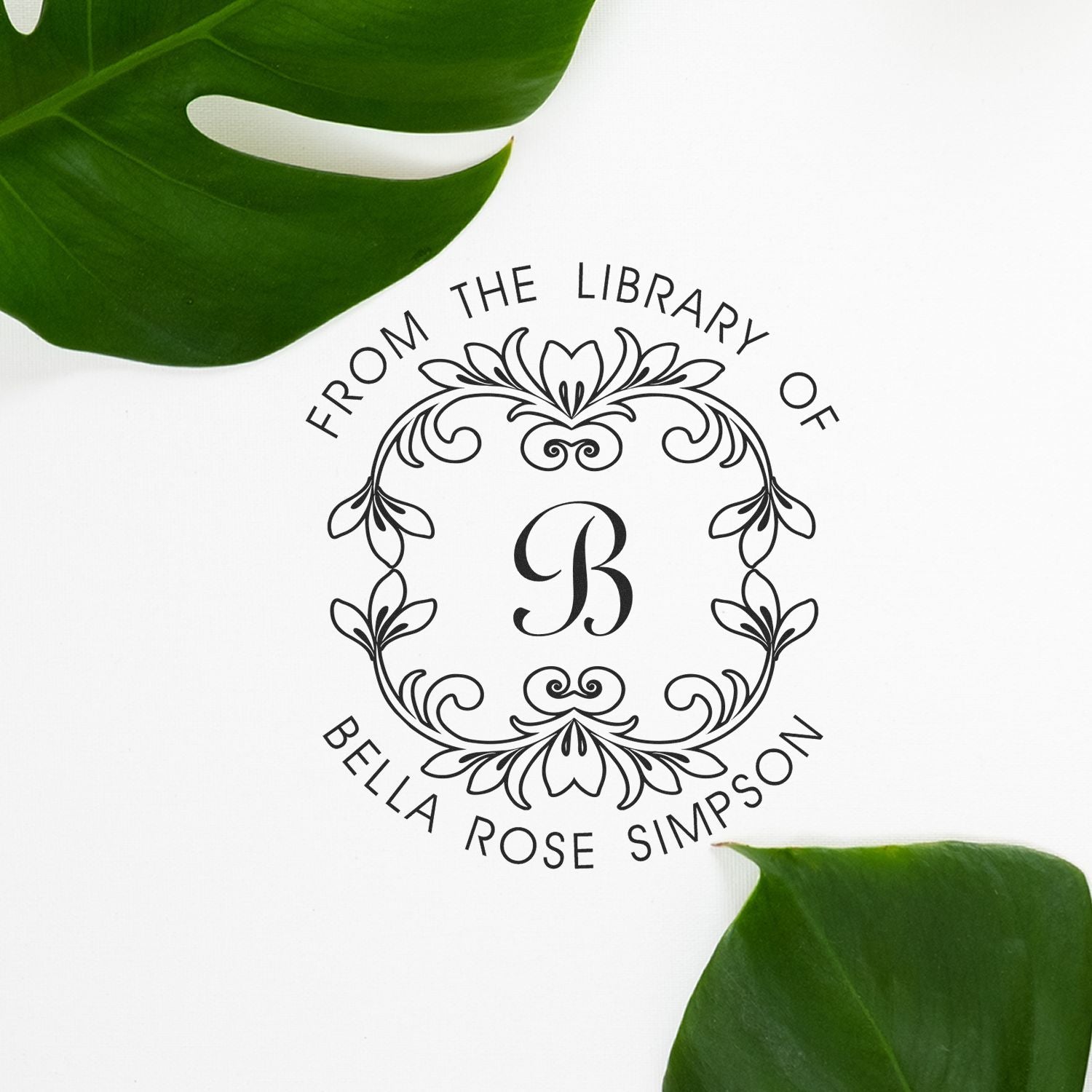 Decorative black library stamp seal with B monogram and floral border, surrounded by green leaves on white background.