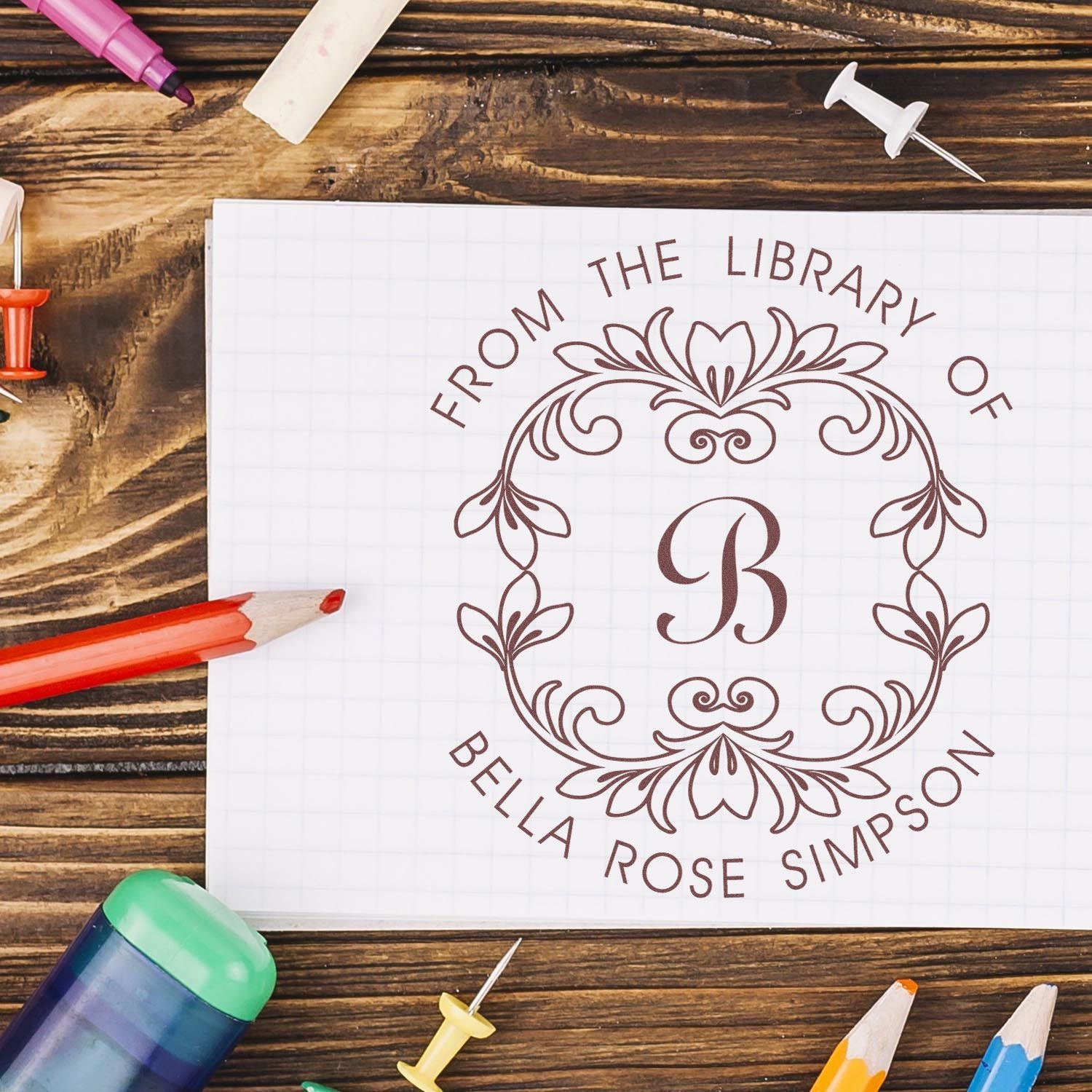 Brown personalized library stamp seal with ornate floral monogram B on white grid paper, surrounded by colorful pencils.