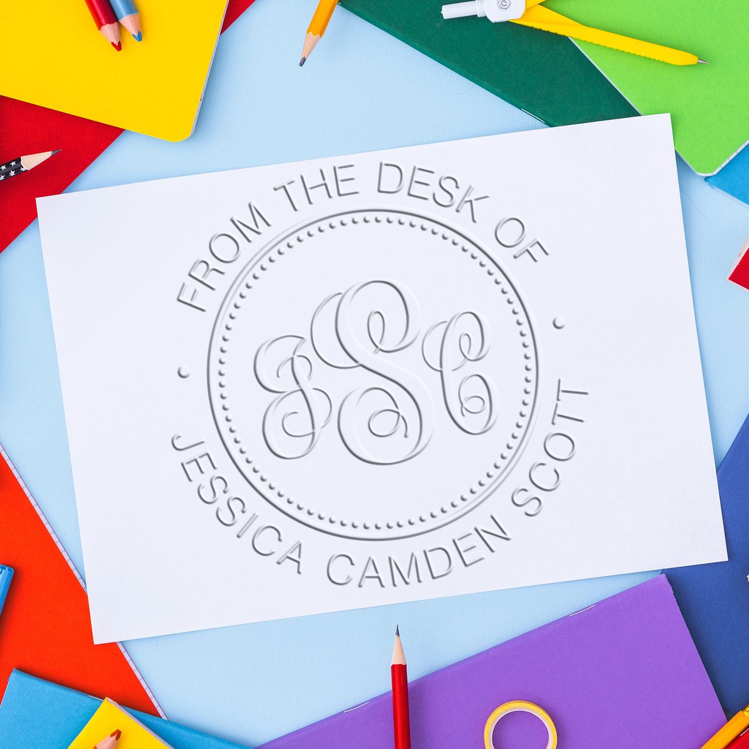 Custom embossed desk seal reading From the Desk of Jessica Camden Scott, on white paper with colorful pencils and notebooks.