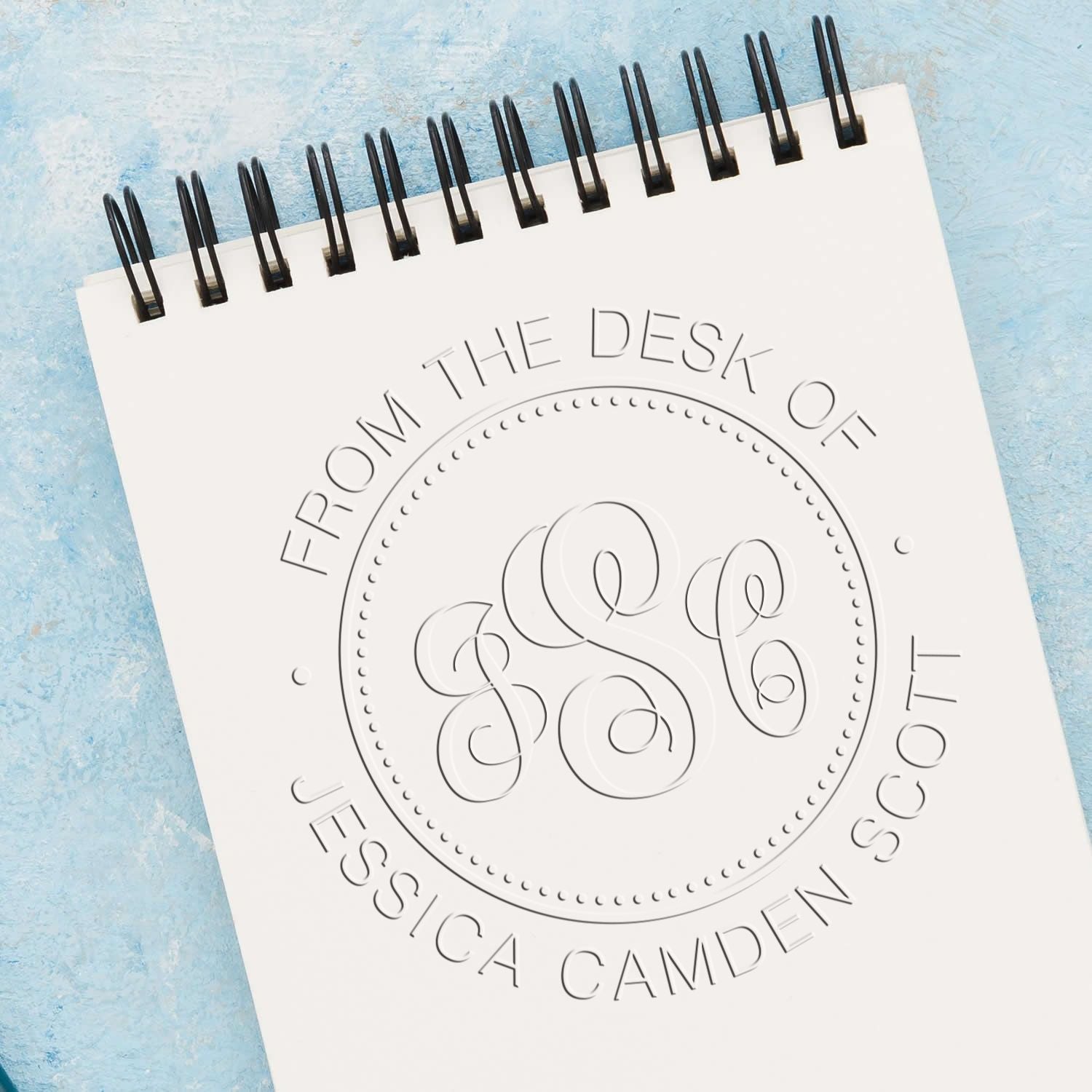 Embossed custom stamp seal on white spiral notebook with black binding, against blue background, reading From the Desk of Jessica Camden Scott