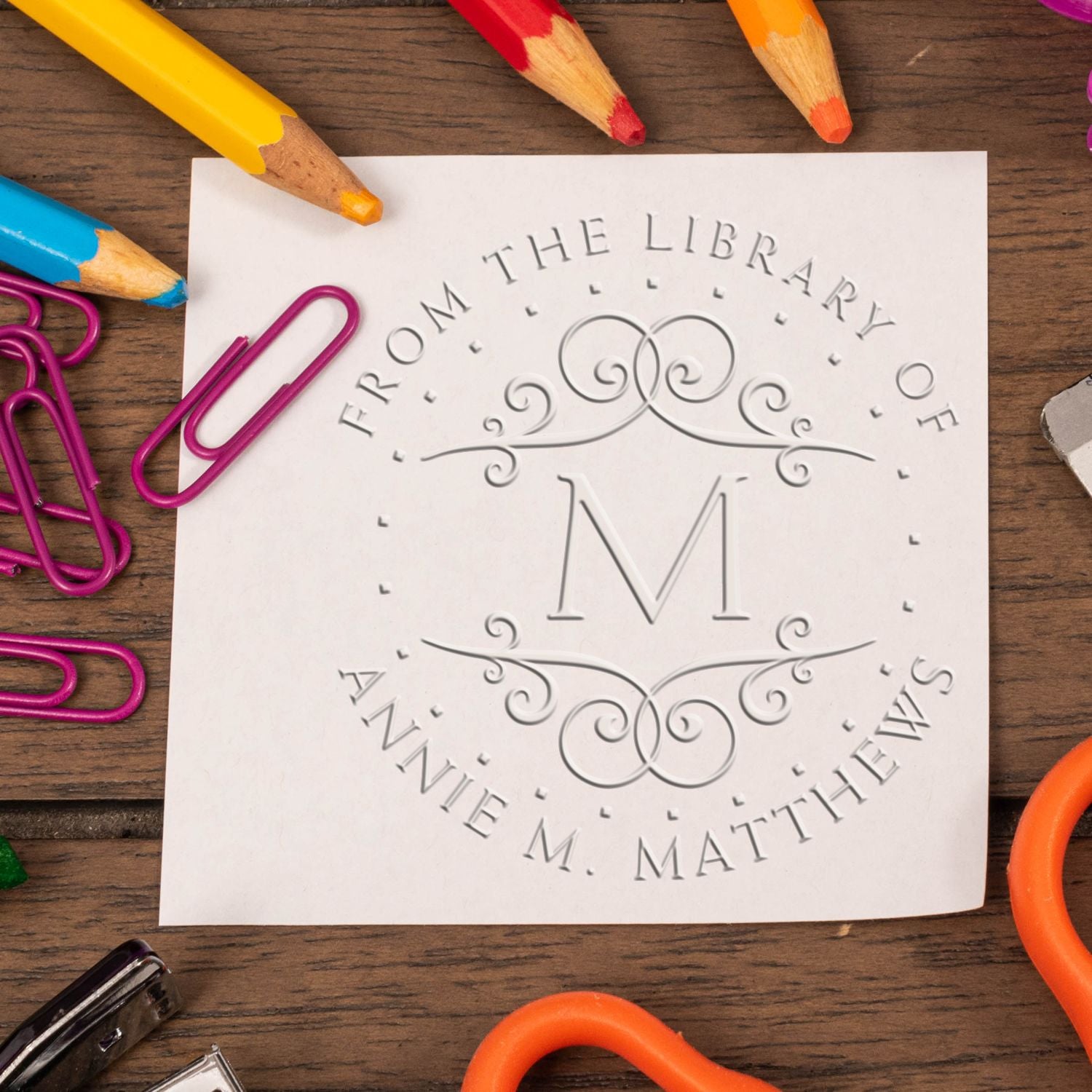 White embossed library stamp seal with monogram M, surrounded by colorful pencils and magenta paper clips on wood.