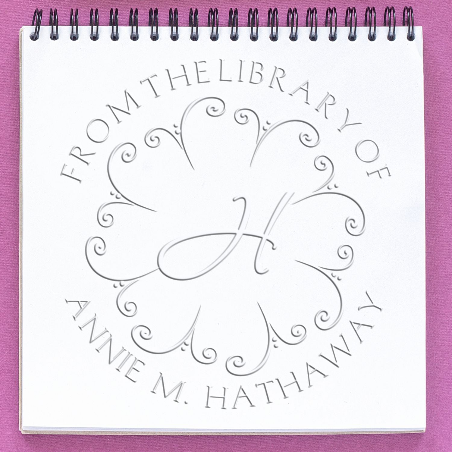 Embossed library seal stamp on white spiral notebook, reading From the Library of Annie M. Hathaway, on a purple background.