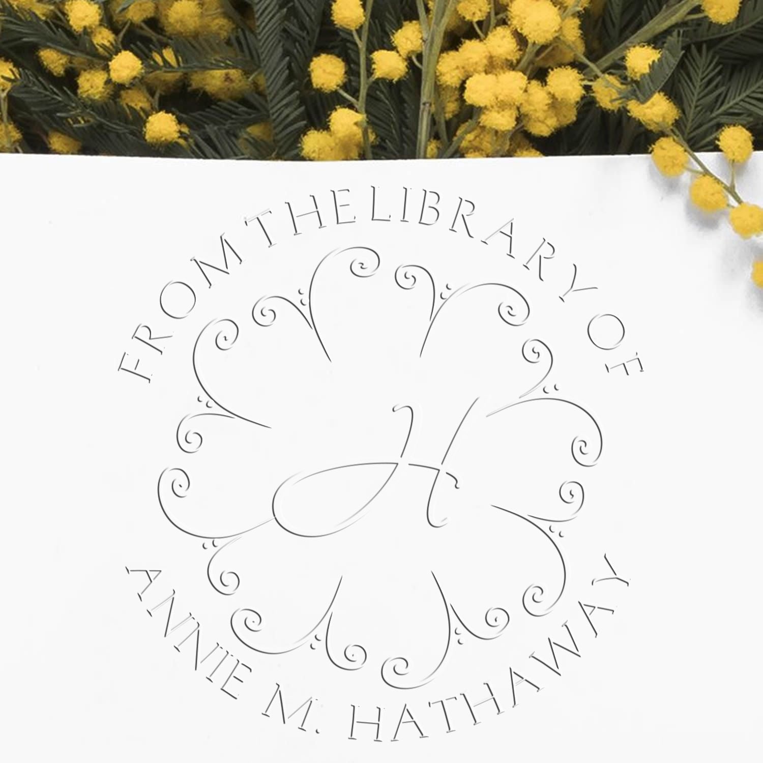 Custom library stamp seal on white, gray floral monogram with From the Library of Annie M. Hathaway, framed by yellow mimosa blooms.