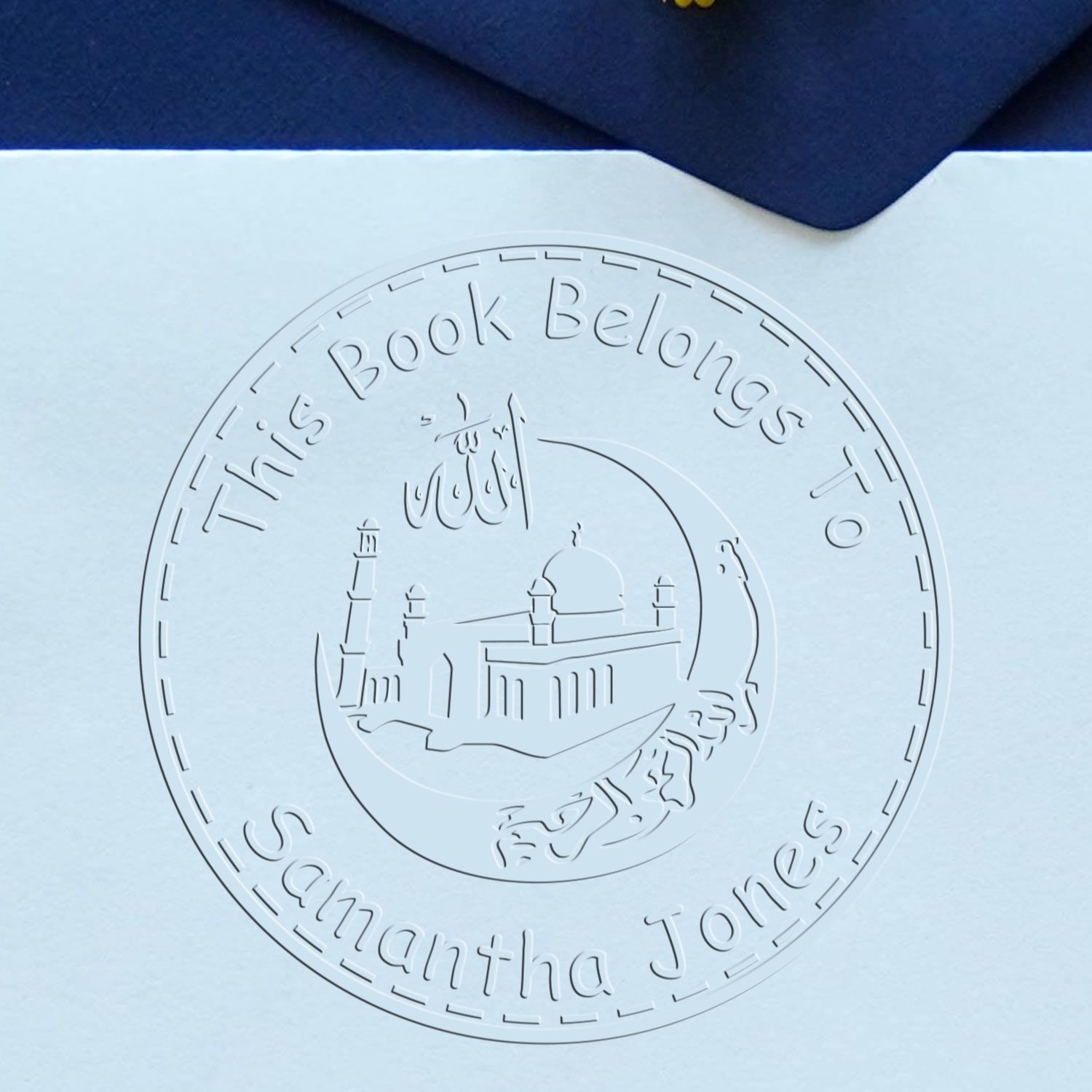 White embossed book stamp seal on light blue paper with navy corner, reading This Book Belongs to Samantha Jones and mosque motif.