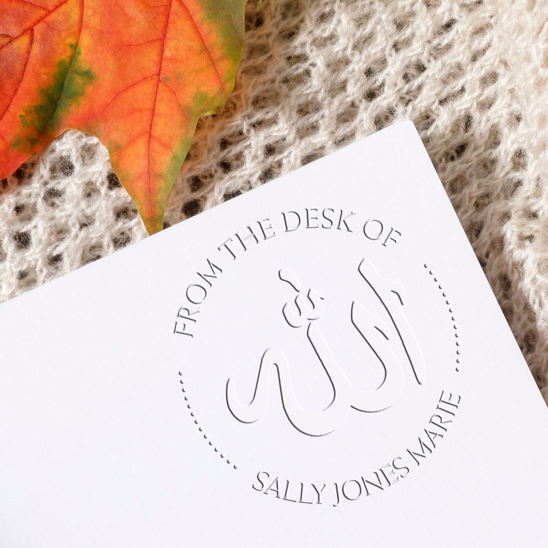 White embossed custom desk stamp and seal reading From the Desk of Sally Jones Marie on a knit background with autumn leaf.
