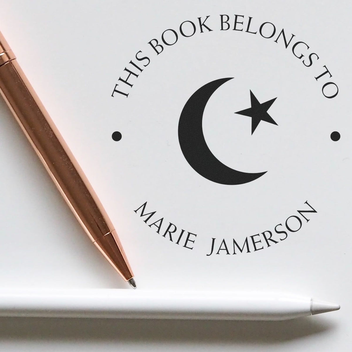 White book stamp and seal with black crescent moon, star, and Marie Jamerson text; rose-gold pen and white stylus beside it.