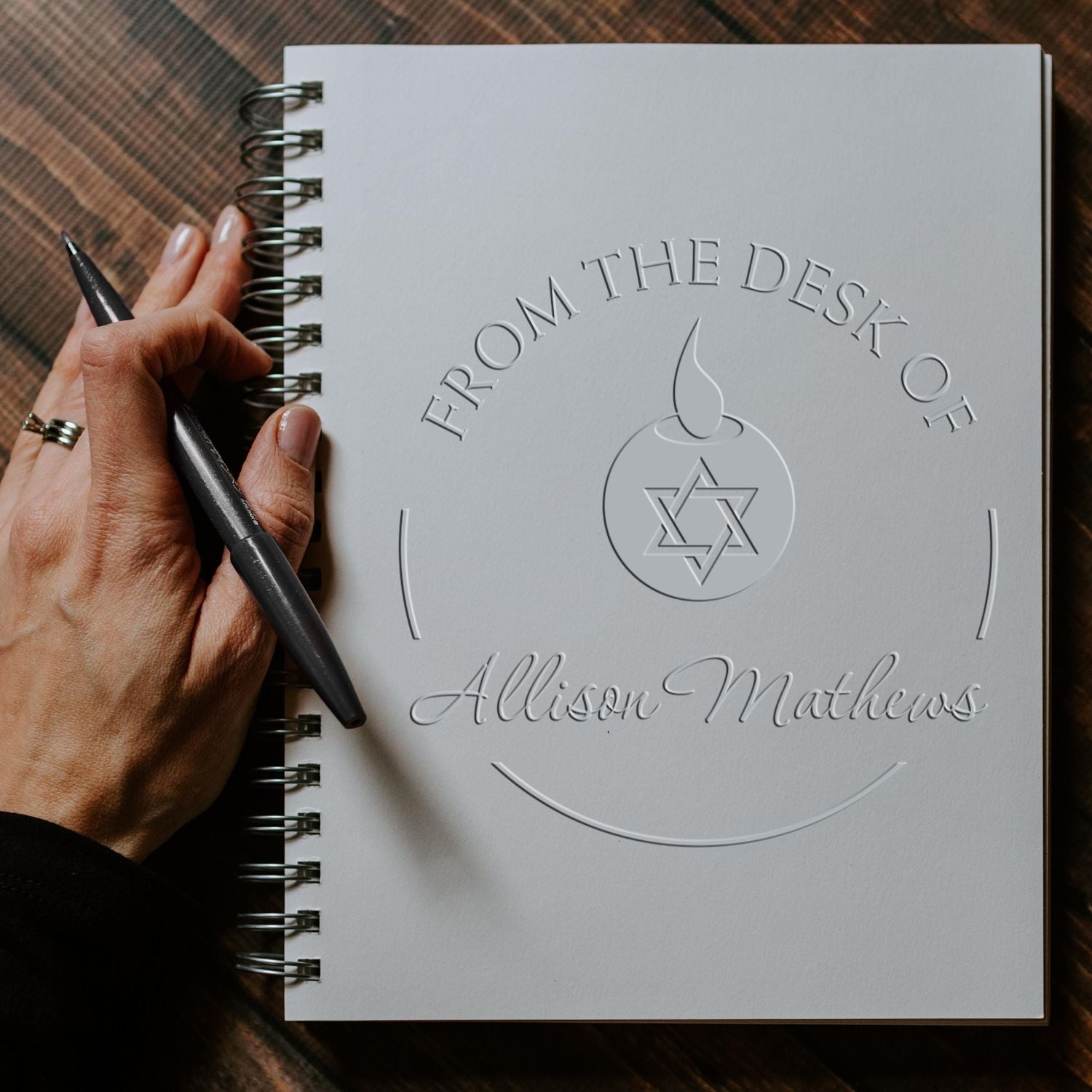 White custom embossed stamp and seal notebook with silver spiral binding, black pen, and hand on a brown textured background.