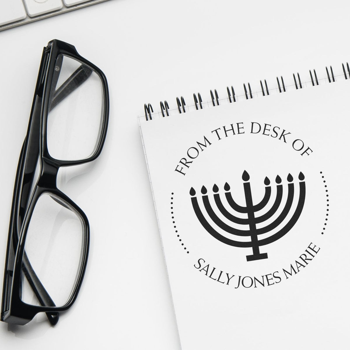 Black custom stamp and seal with menorah design and From the Desk of Sally Jones Marie beside black eyeglasses on white background.