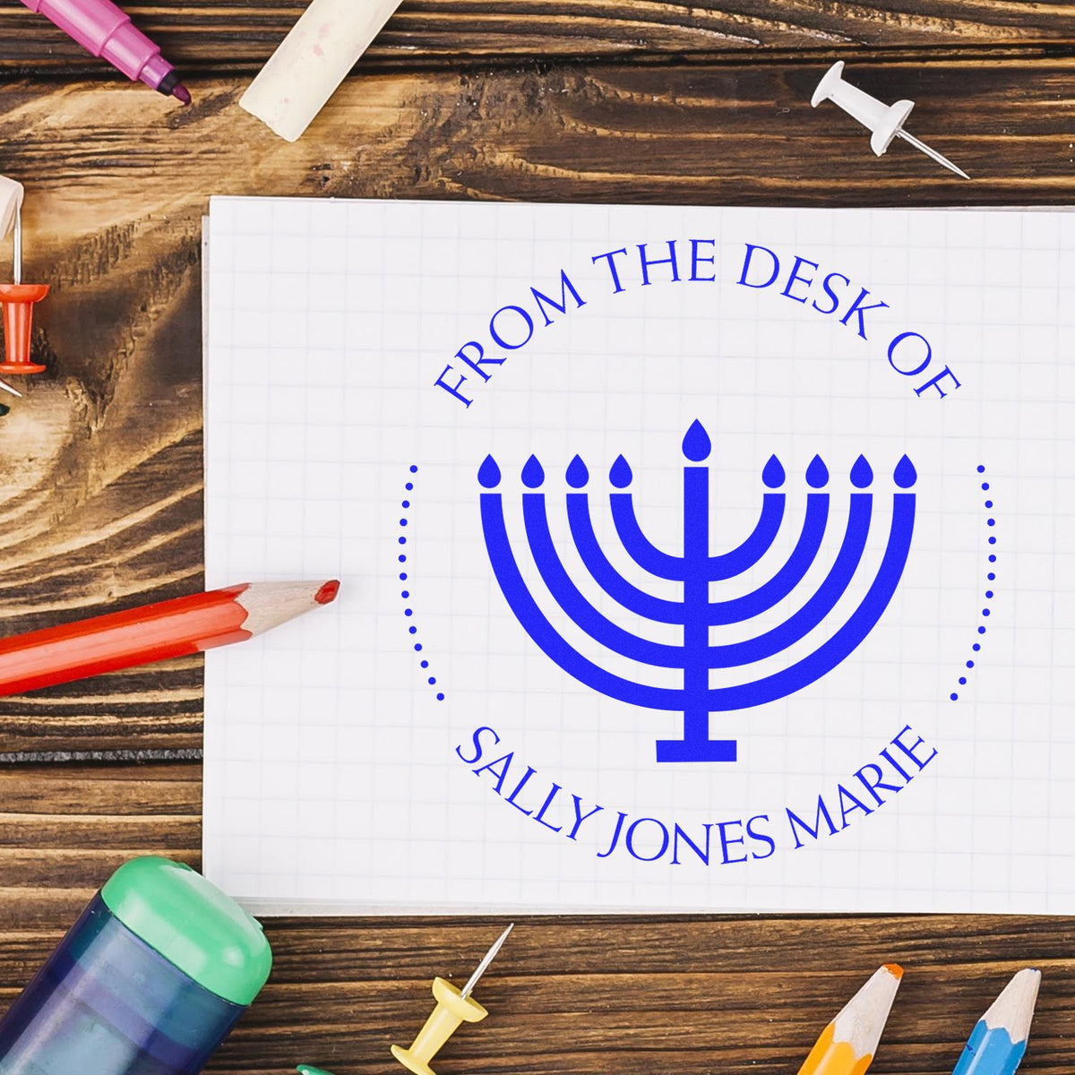 Blue menorah stamp and seal on white grid paper, with colored pencils and pushpins on a rustic wood desk.