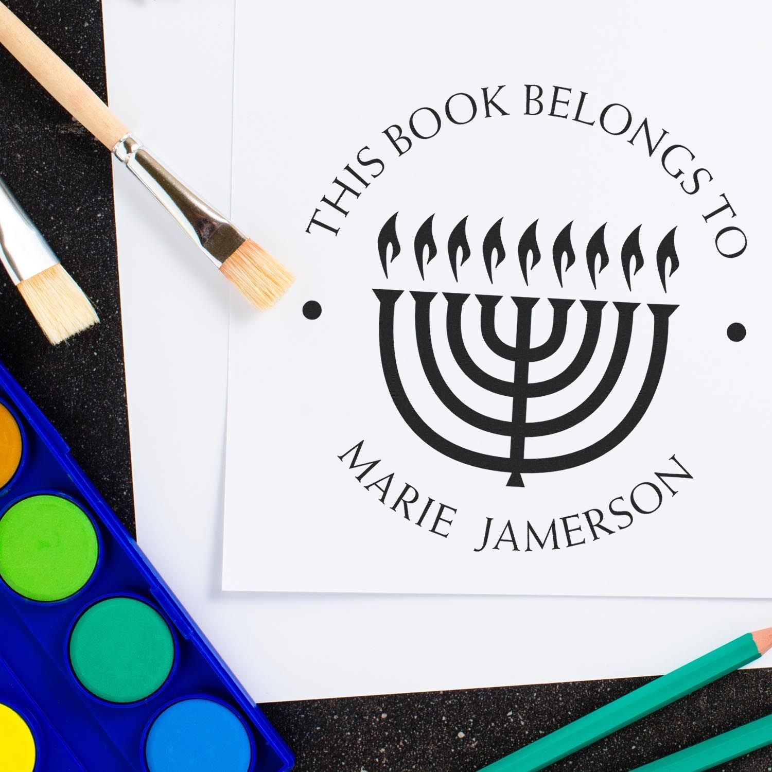 Custom black book stamp and seal featuring a menorah and This Book Belongs to Marie Jamerson, with brushes and bright paints.