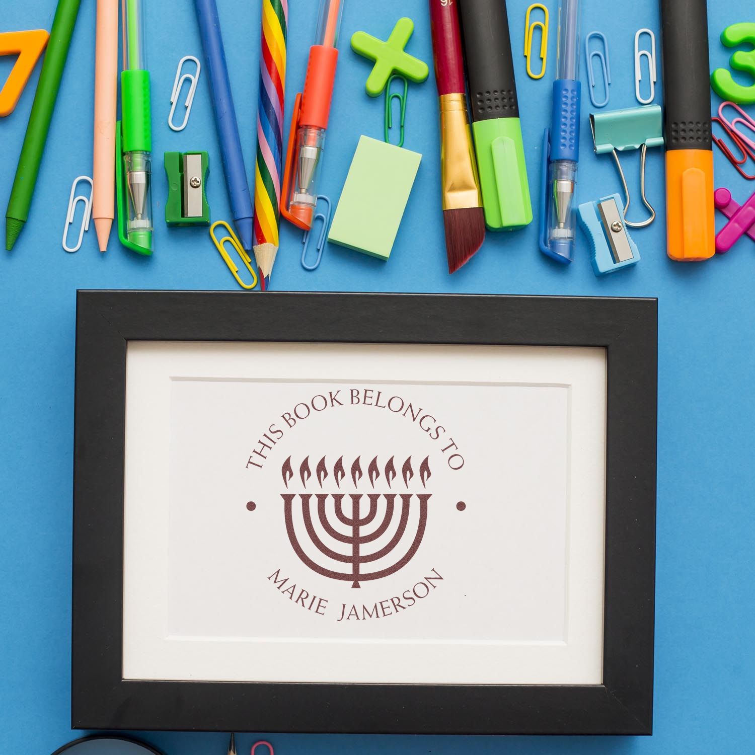 Colorful stationery on blue background with a framed maroon menorah bookplate stamp and seal reading This Book Belongs to Marie Jamerson.