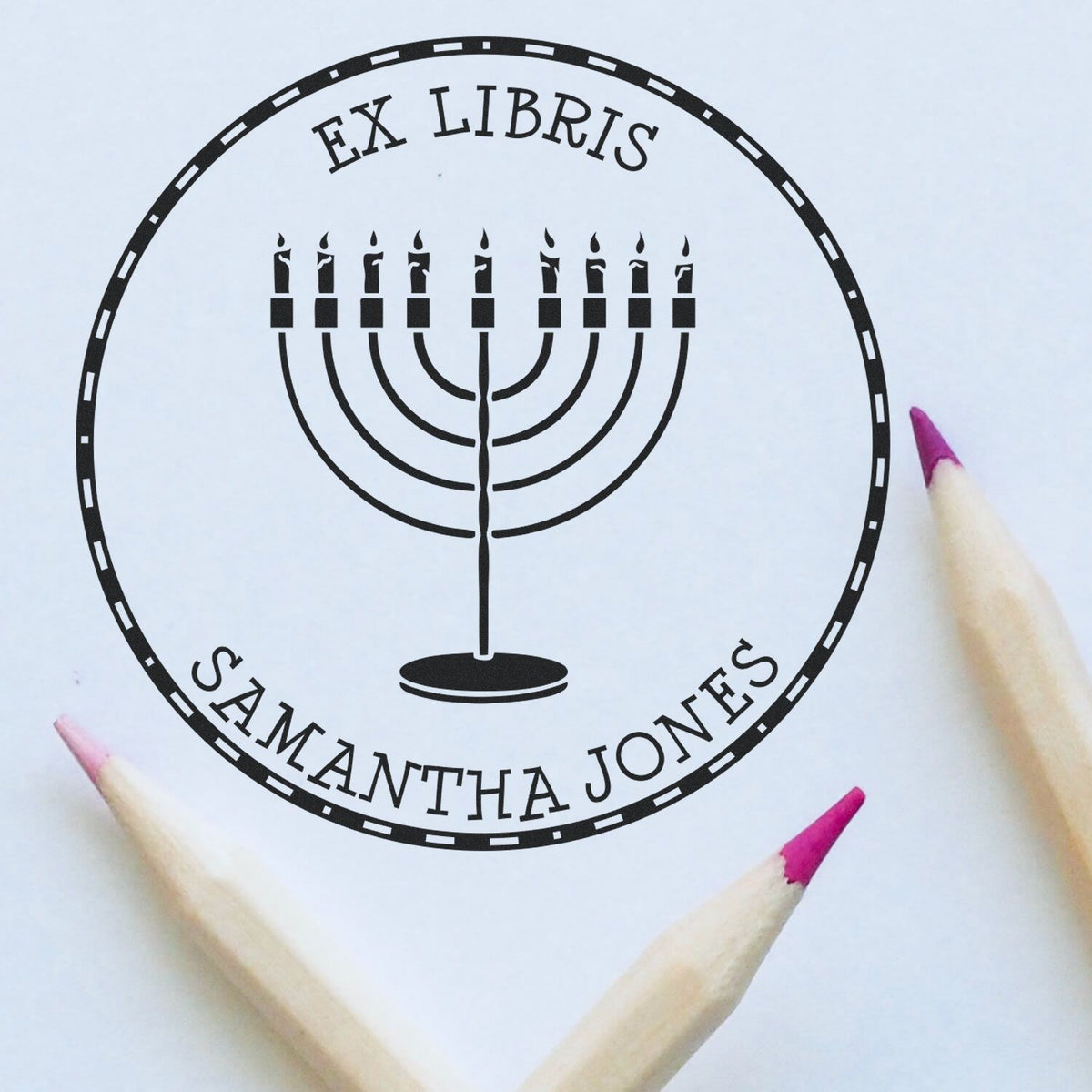 Black circular ex libris stamp seal with menorah and EX LIBRIS SAMANTHA JONES on white paper, framed by pink pencil tips.