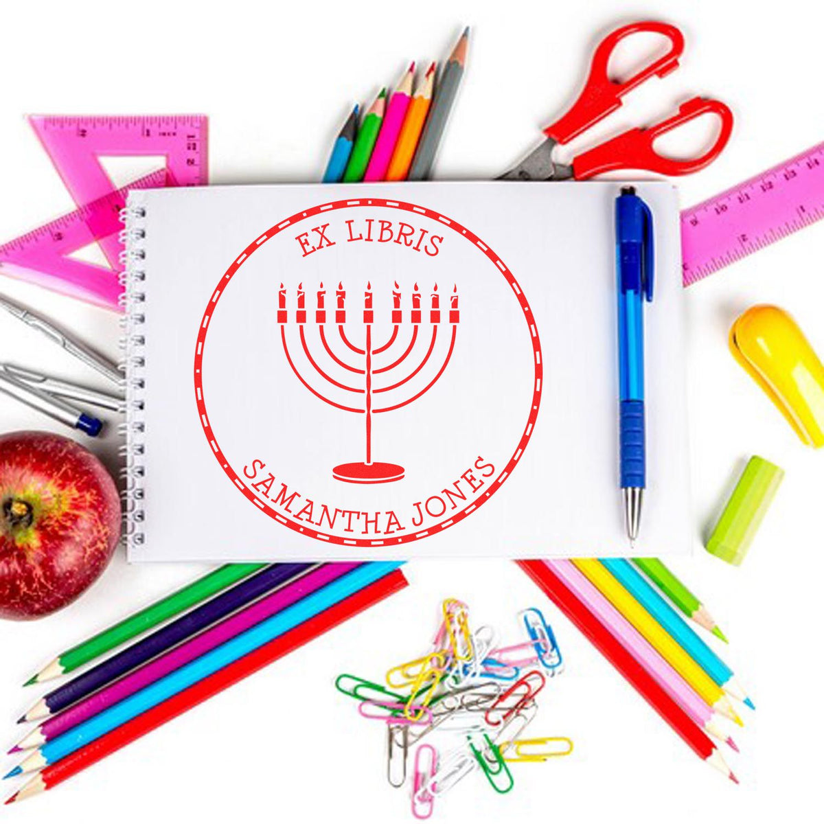 Red custom ex libris stamp seal with menorah on notebook, surrounded by colorful pencils, rulers, pens, scissors, and paper clips.