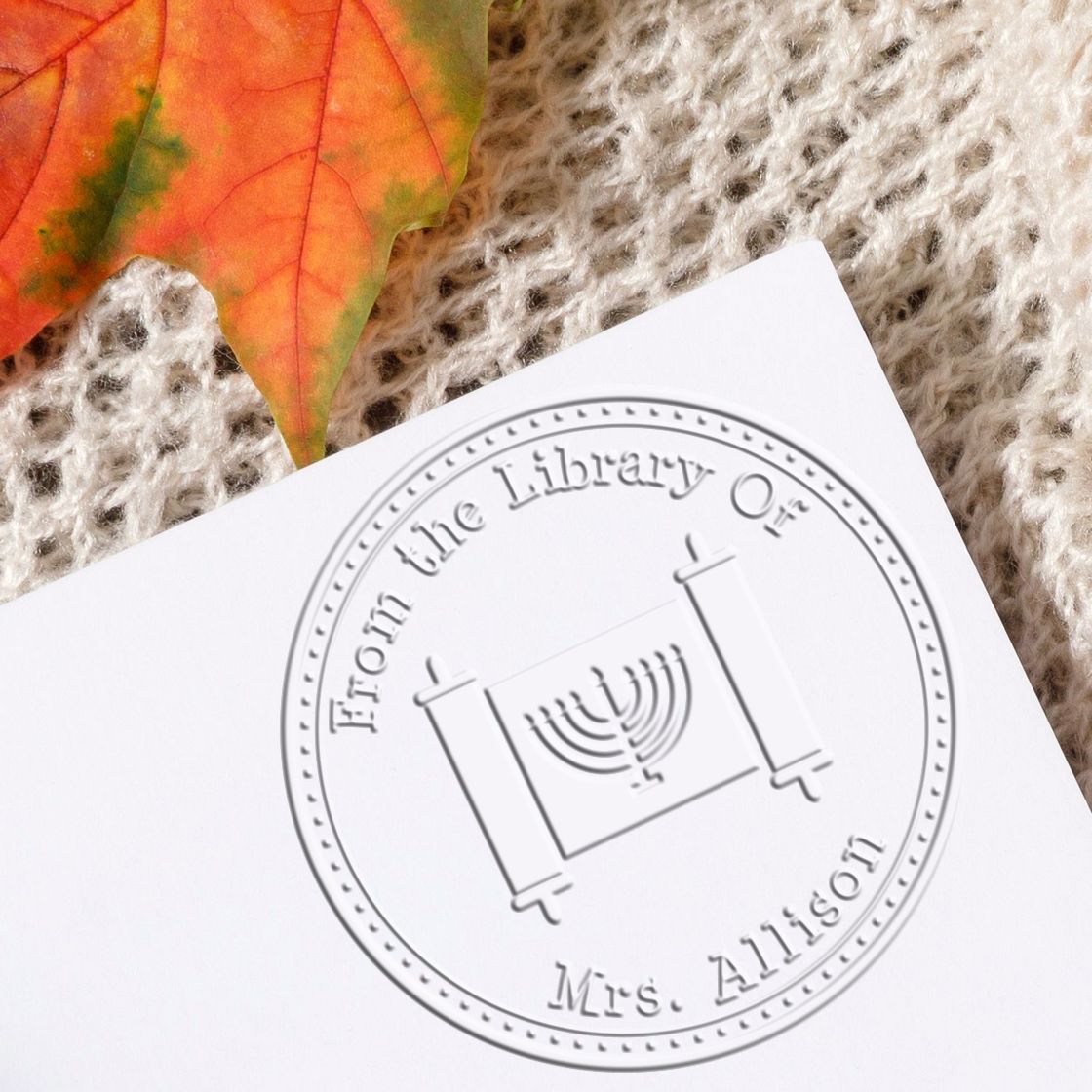 Embossed white library stamp seal on paper, reading From the Library of Mrs. Allison, with a menorah emblem and orange leaf.