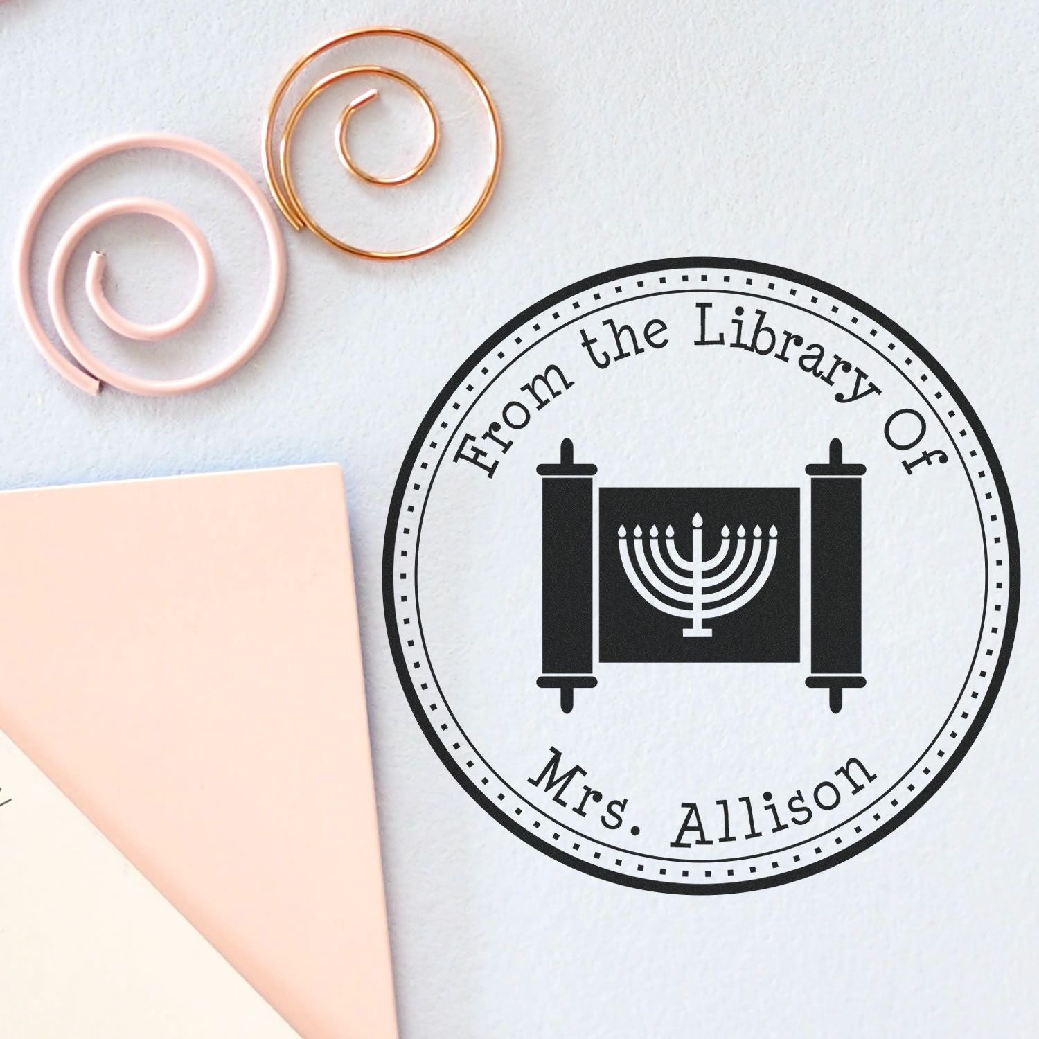 Black custom library stamp seal with menorah, white background, pale pink bookmark, and pink-gold spiral paper clips.