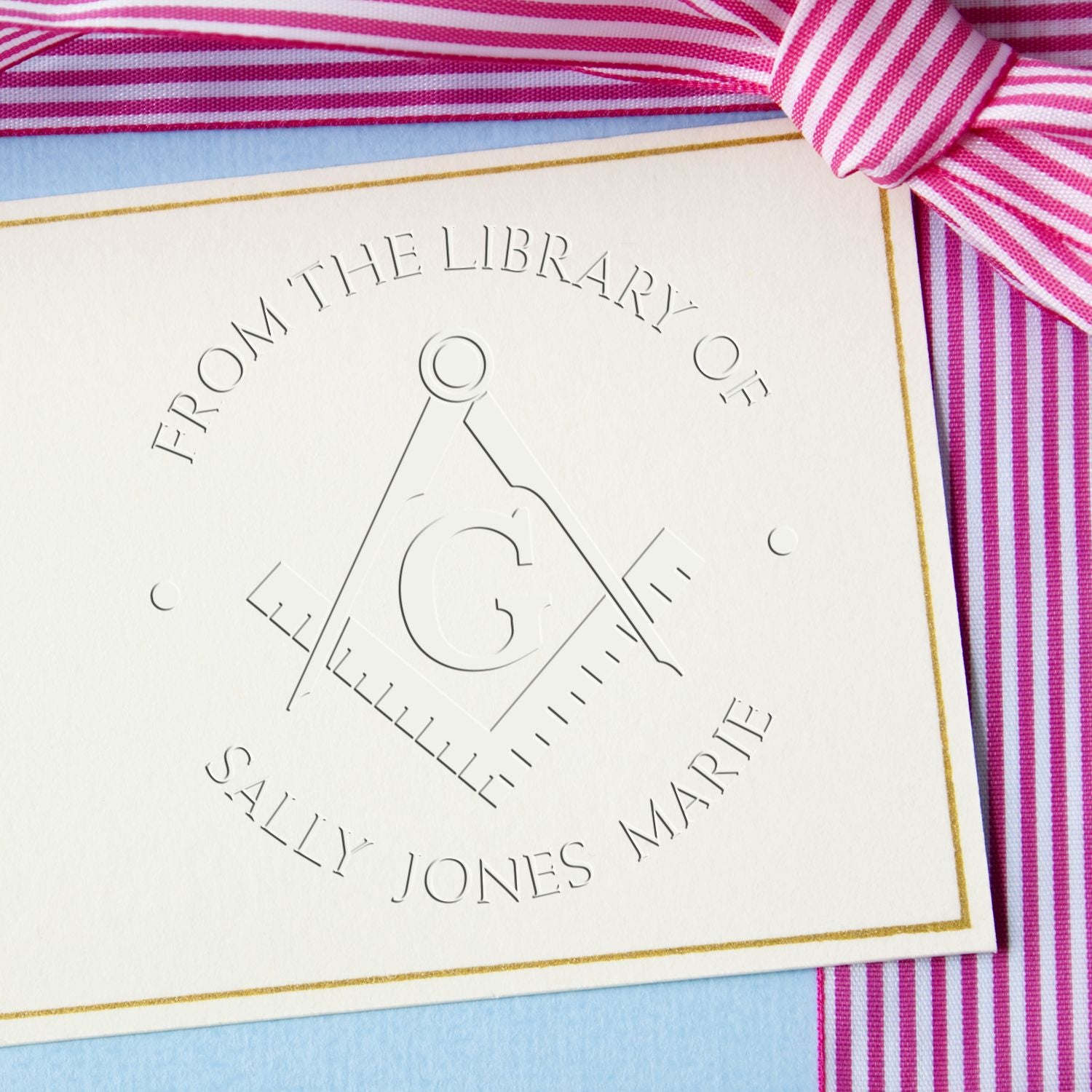 Embossed custom bookplate seal with monogram G on white card, gold border, pink striped ribbon, and blue background.