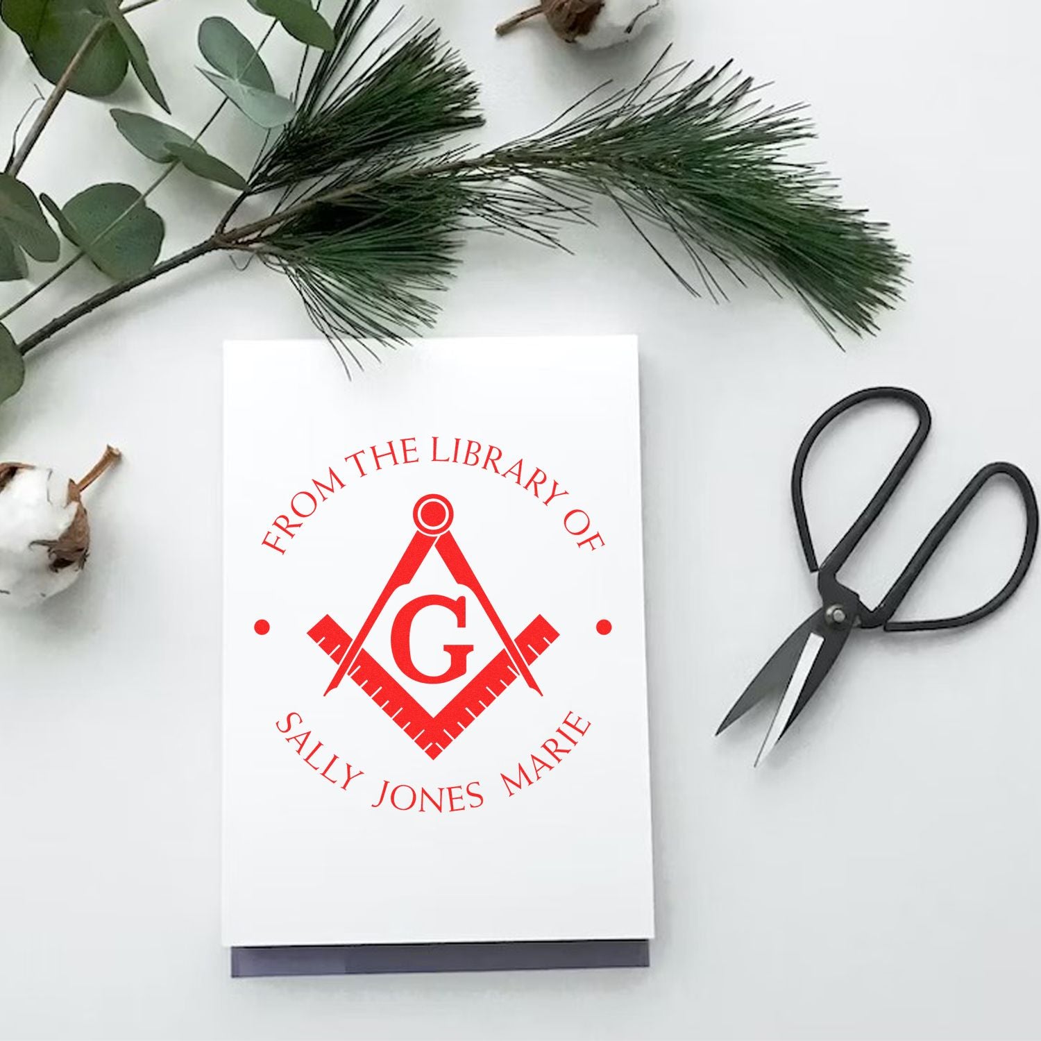 Red personalized library stamp seal on a white card, with black scissors and green foliage.