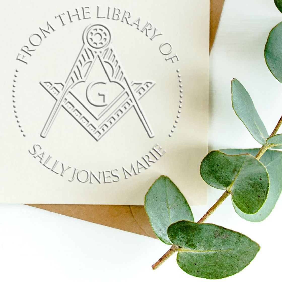 Cream custom library stamp seal with gray Masonic emblem and Sally Jones Marie text, beside eucalyptus leaves and brown envelope.