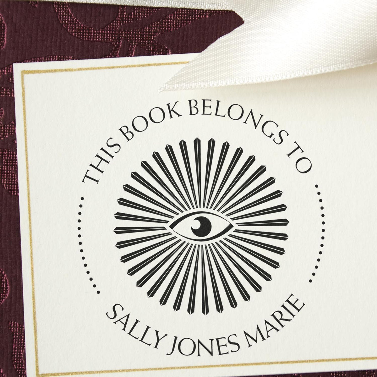 Black bookplate stamp and seal with eye emblem on cream card, reading This book belongs to Sally Jones Marie, on burgundy fabric.