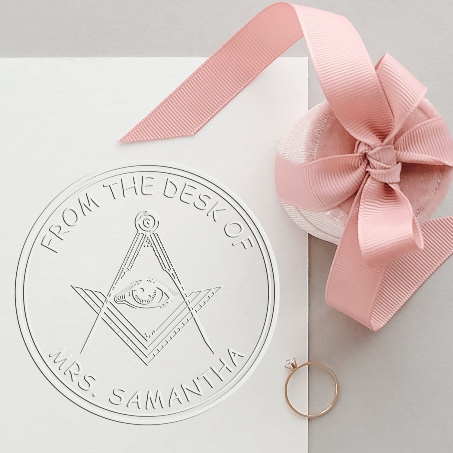 White custom stamp and seal with eye emblem, pink ribbon bow, and gold ring on a clean gift package.