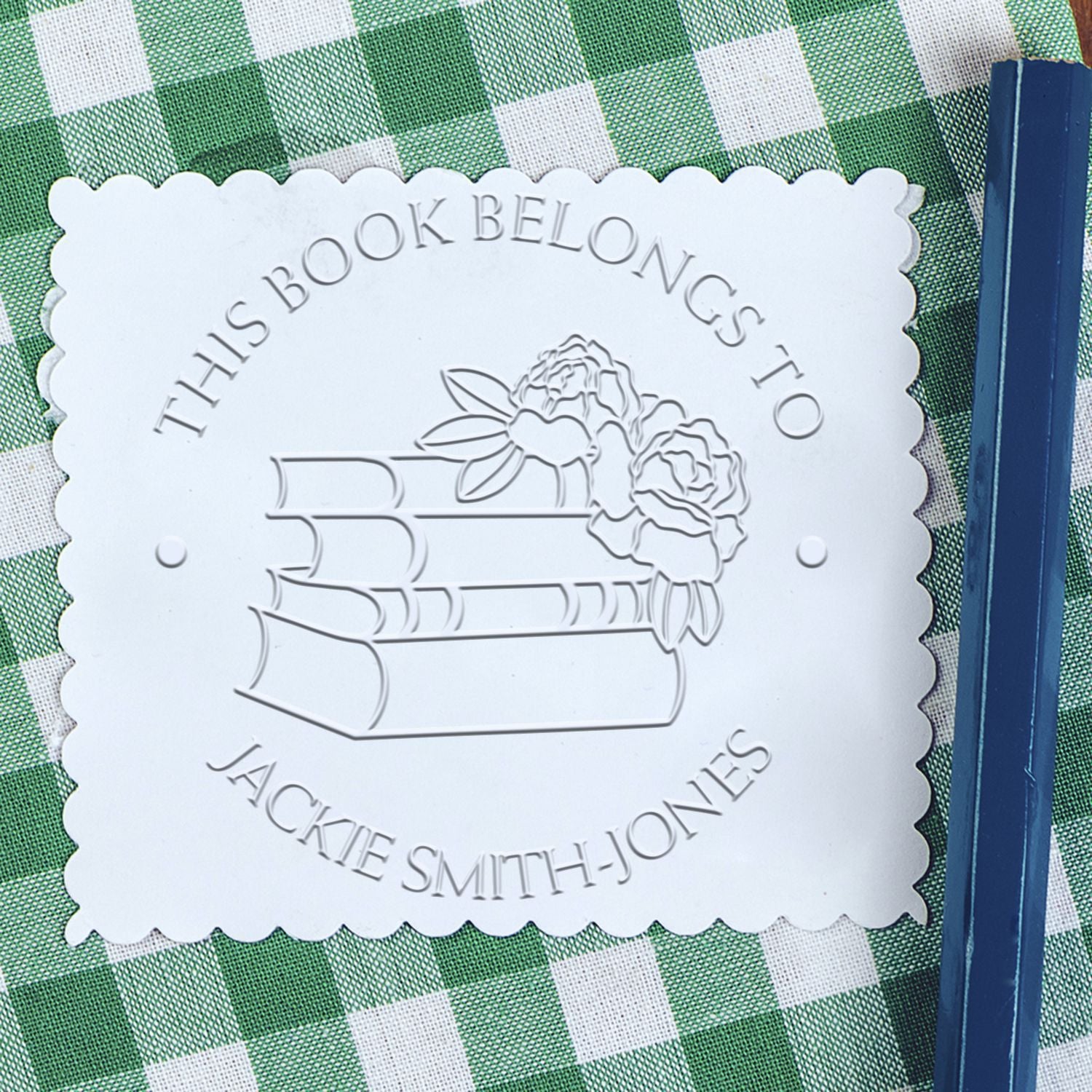 White scalloped embossed bookplate seal with stacked books and flowers on green gingham, beside a blue book spine.