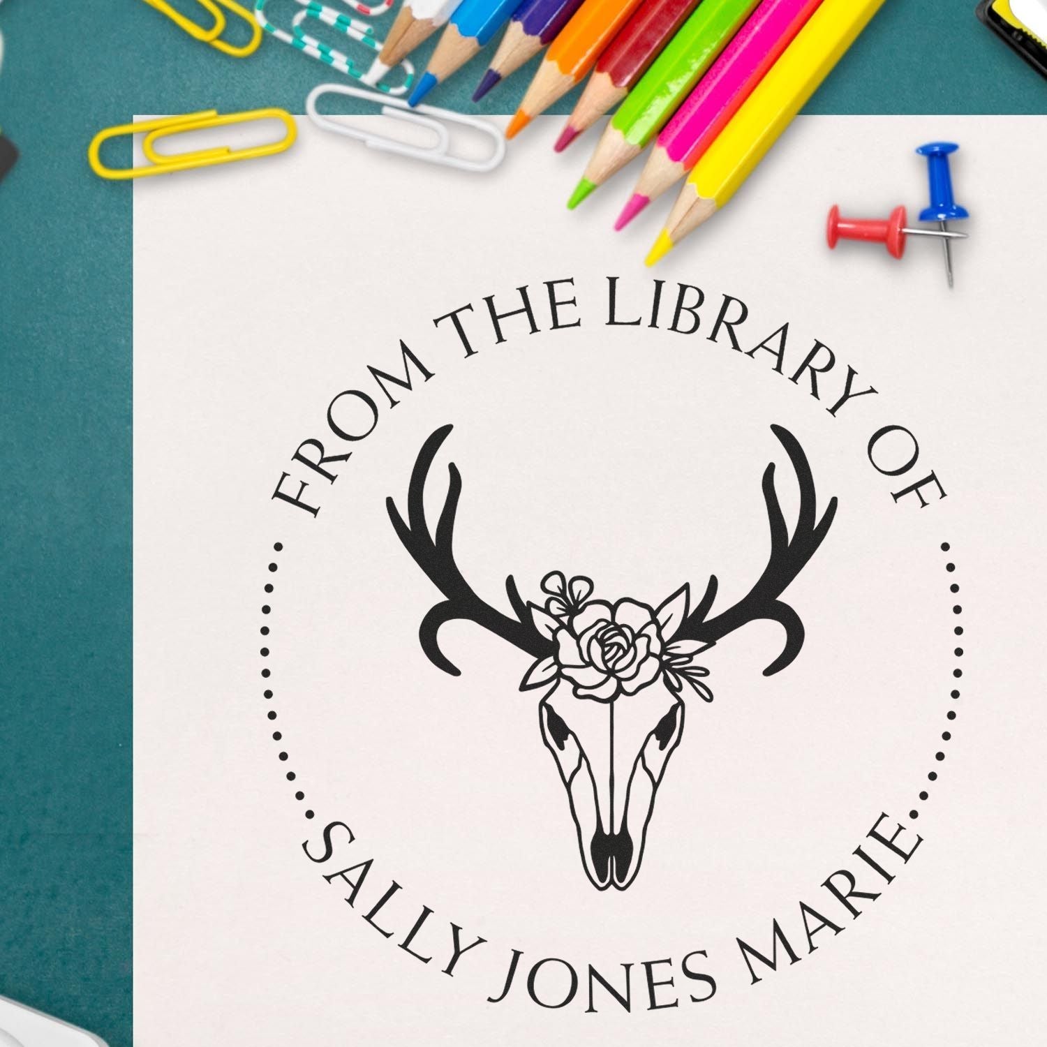 Custom black library stamp and seal with deer skull and floral design, framed by colorful pencils and paper clips.