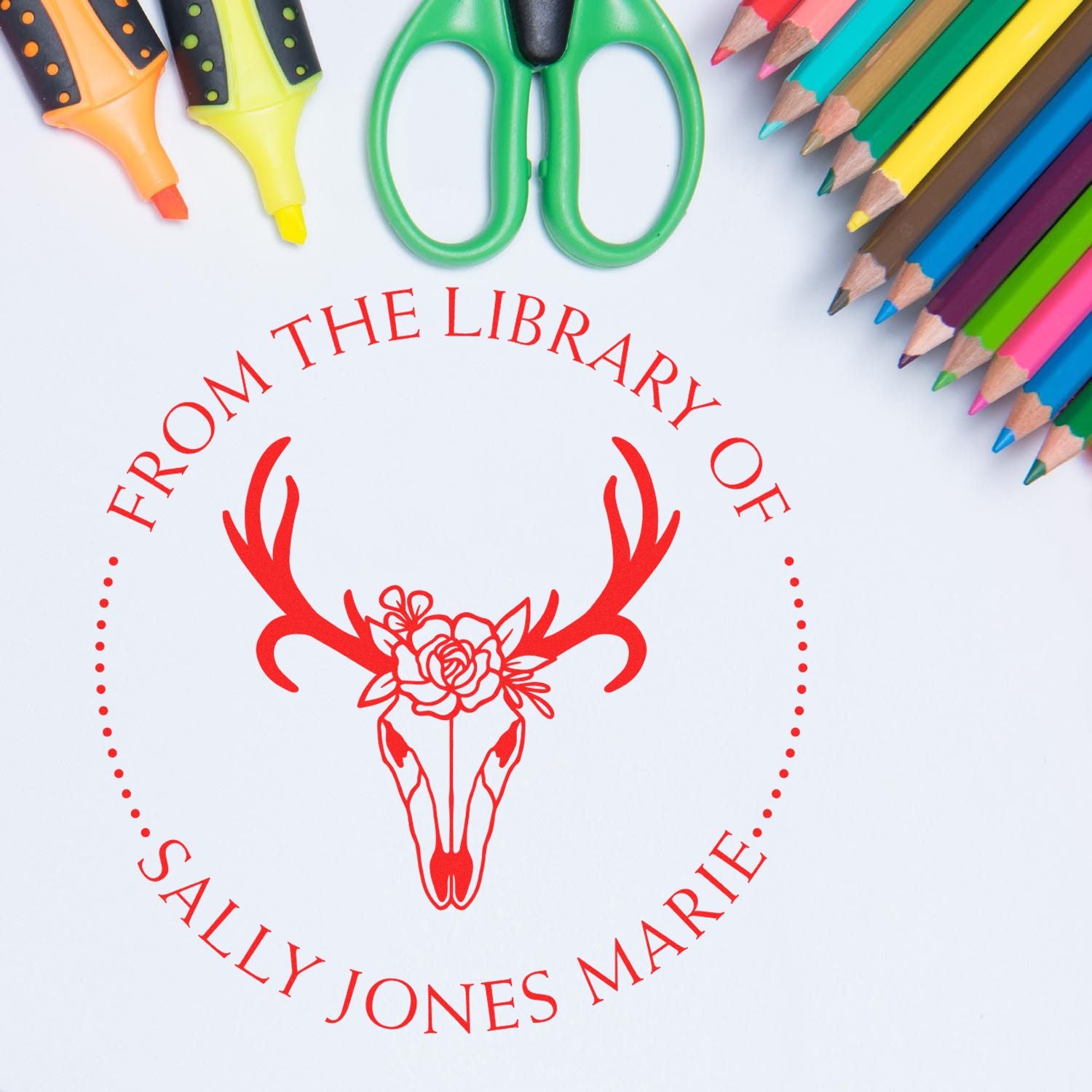 Red custom library stamp seal with deer skull and flowers, surrounded by colorful pencils, markers, and green scissors.