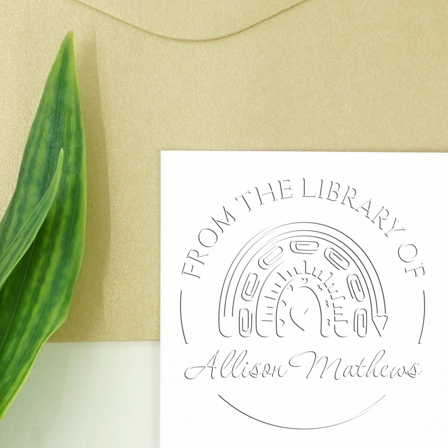 White custom library seal stamp with embossed rainbow design and Allison Mathews text on gold background, beside green leaf