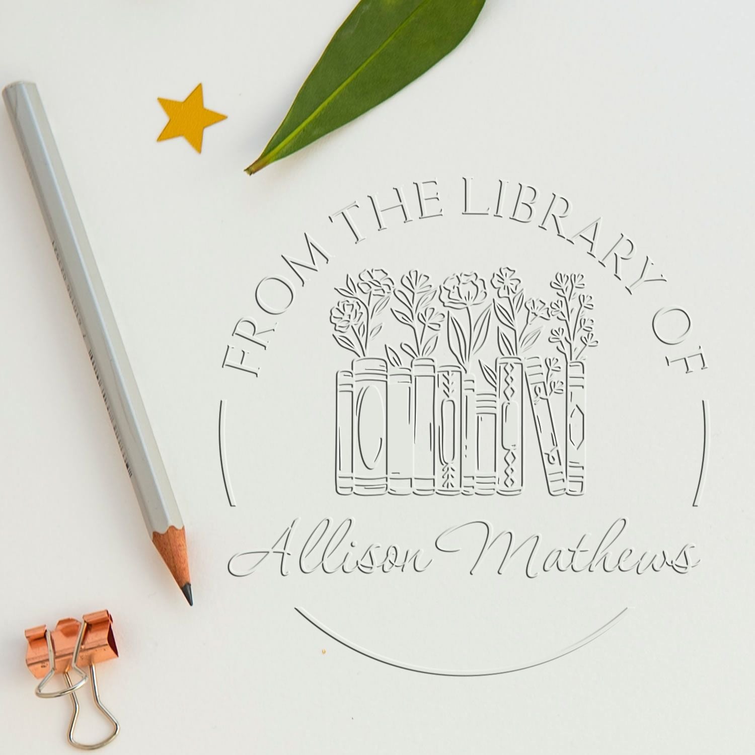 Clear custom library stamp and seal with engraved books and flowers, beside a gray pencil, green leaf, and gold star.