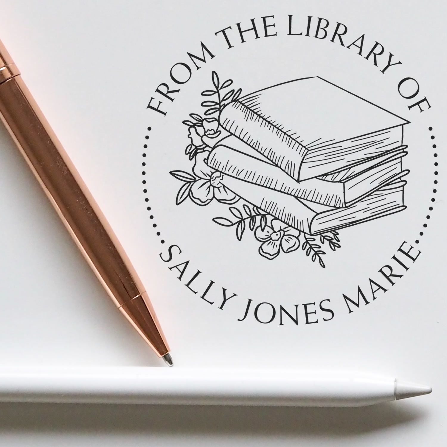 Custom library book stamp and seal with stacked books and flowers, black on white, beside a rose-gold pen and white stylus.