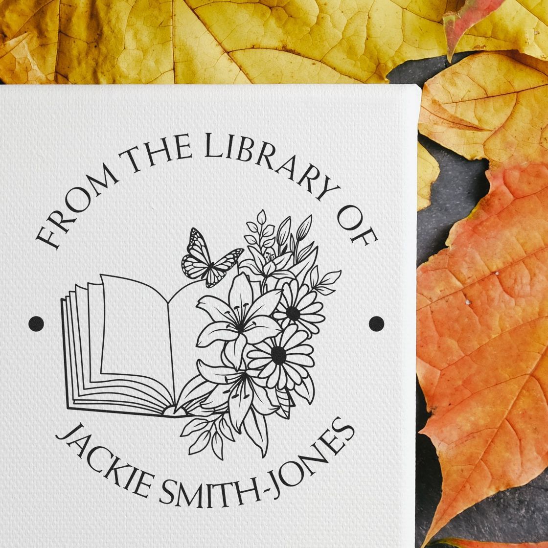 Black custom library stamp seal with open book, flowers and butterfly on white paper, surrounded by yellow-orange autumn leaves.