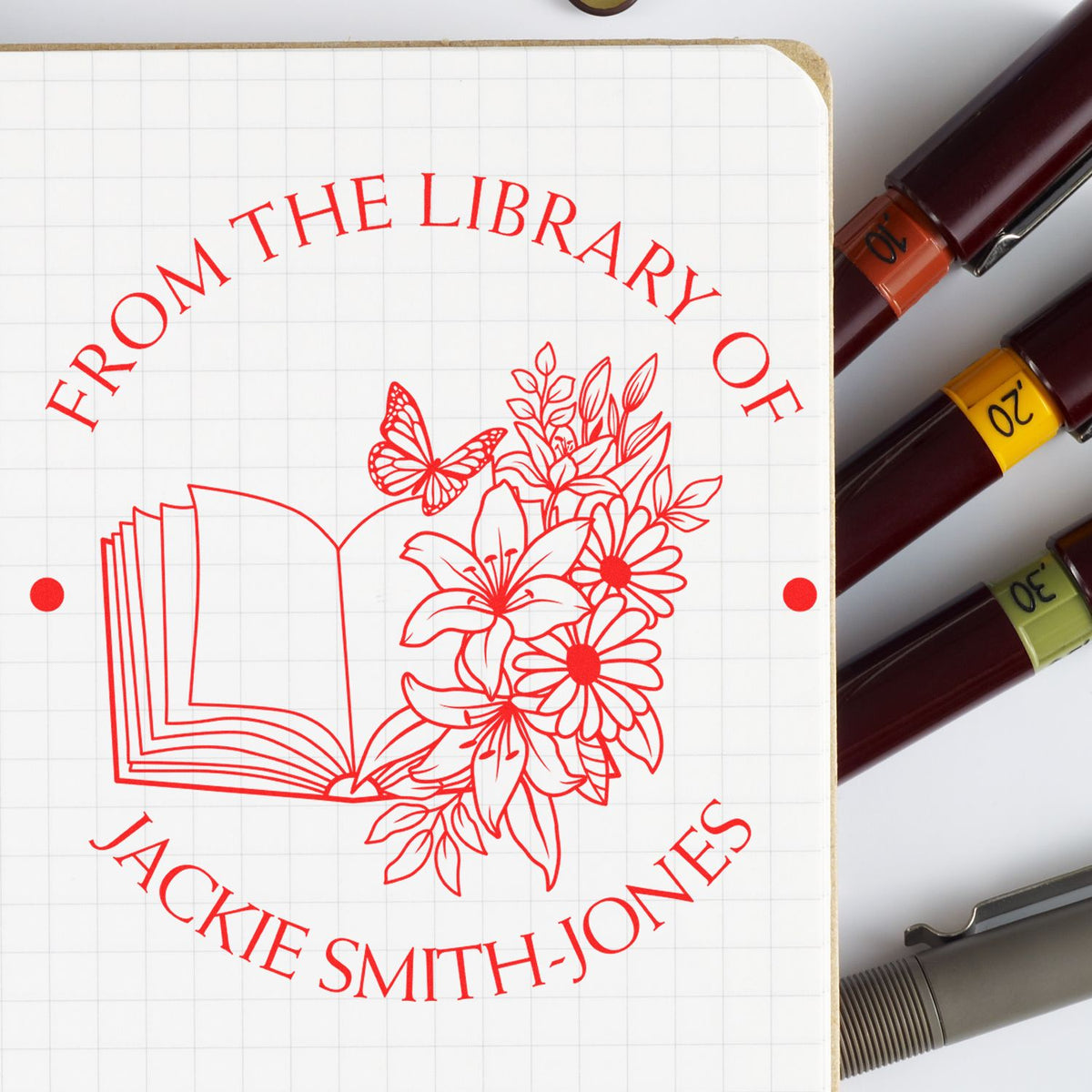 Red library stamp seal with book, flowers and butterfly on grid paper, beside burgundy, yellow, green and gray stamp pens.