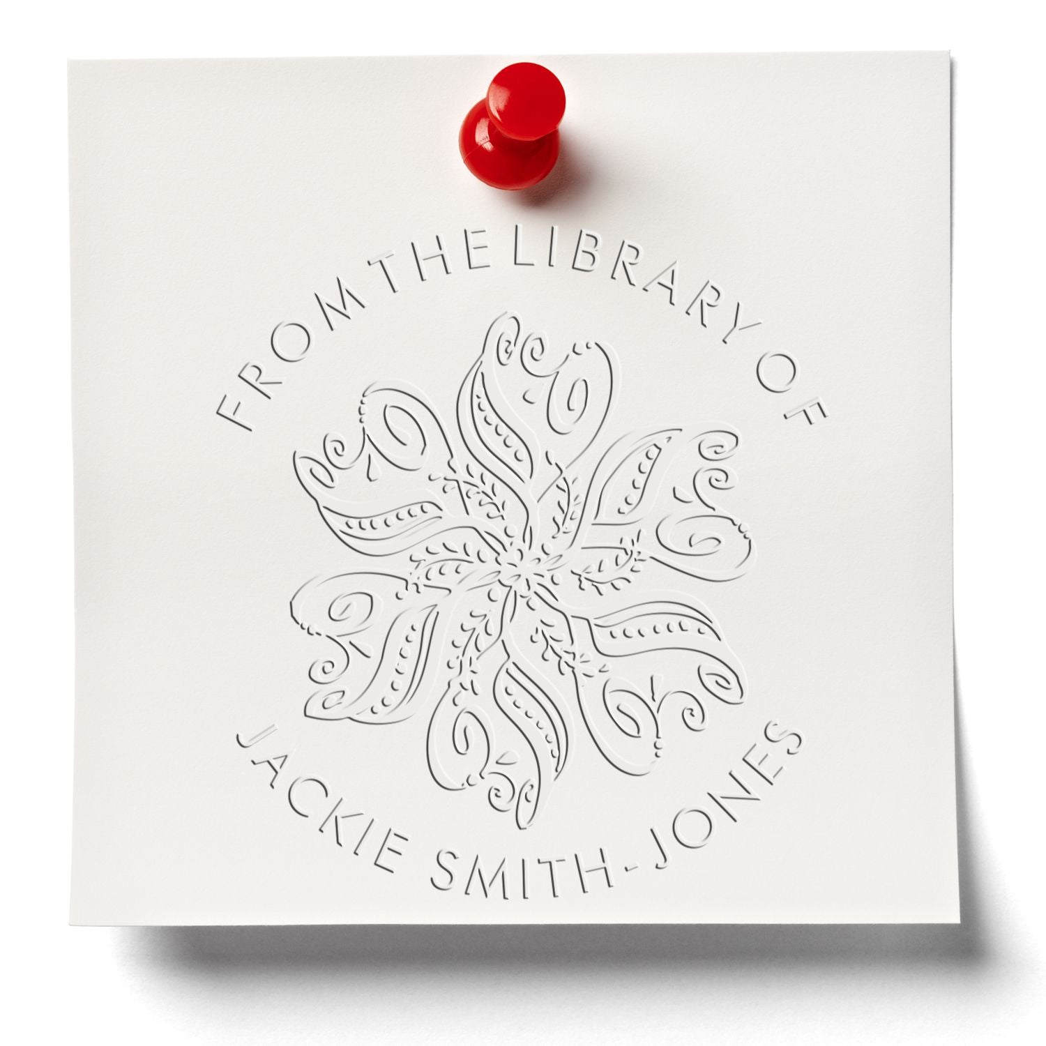 White embossed library seal stamp with ornate floral motif and red pushpin, reading From the Library of Jackie Smith-Jones.