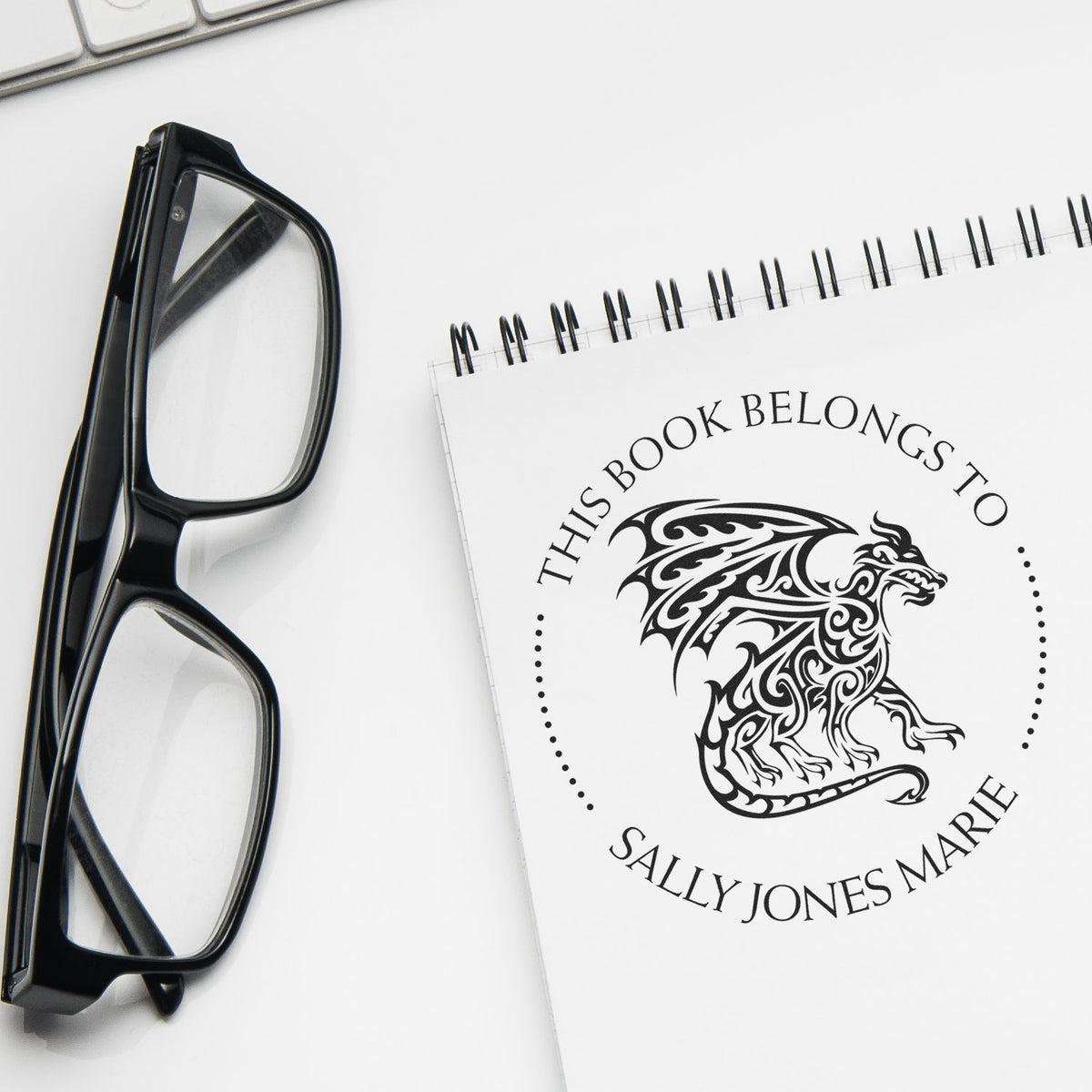 Black custom bookplate stamp and seal with dragon design on a white notebook beside black eyeglasses.
