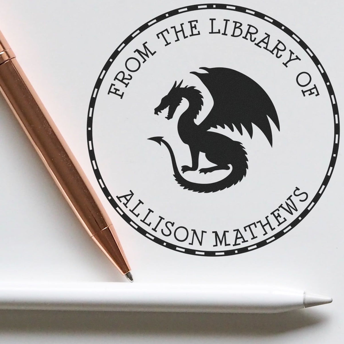 Black circular library stamp seal with dragon emblem and From the Library of Allison Mathews, beside a rose gold pen on white.