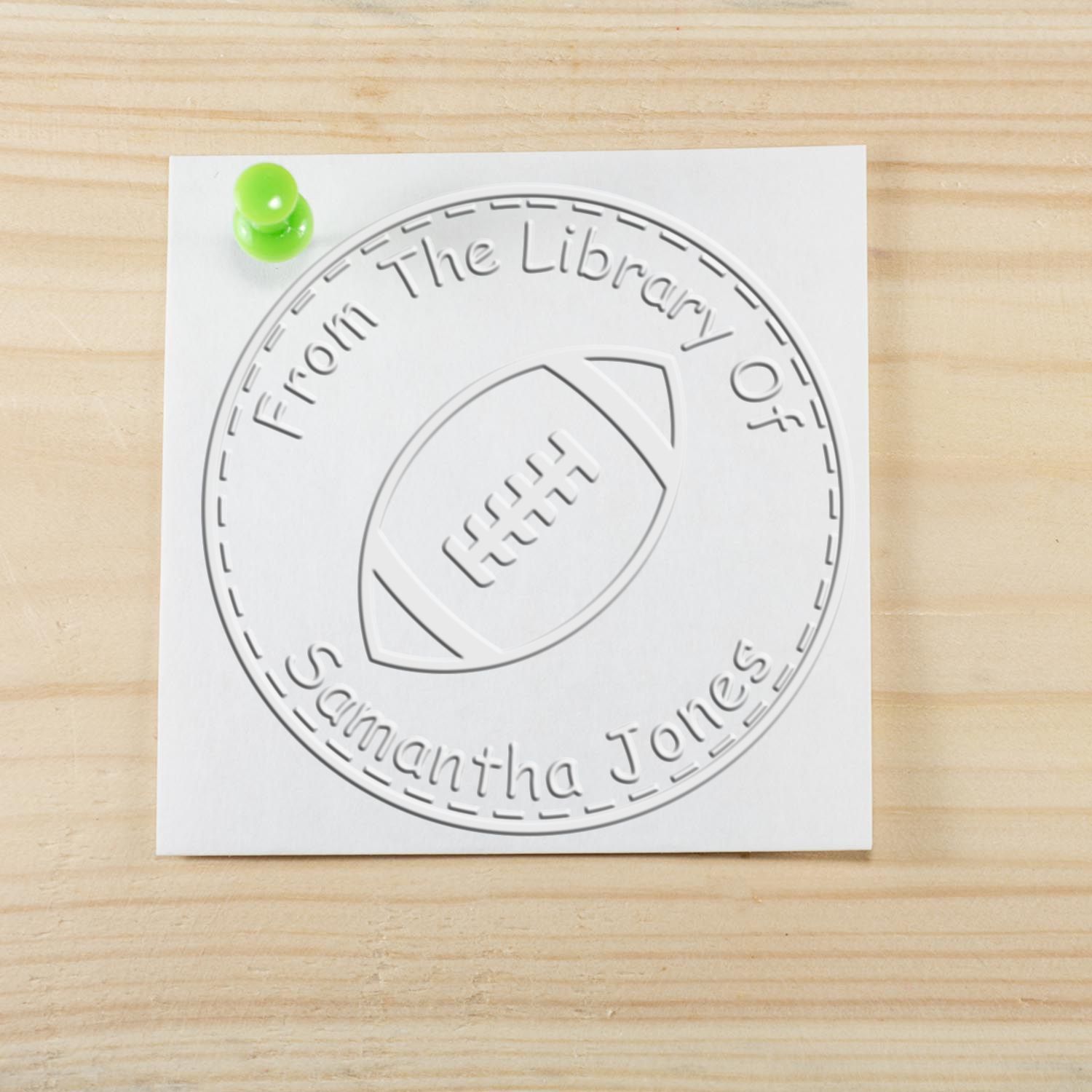 Embossed white library stamp seal with football, From the Library of Samantha Jones, pinned with a green tack on wood.