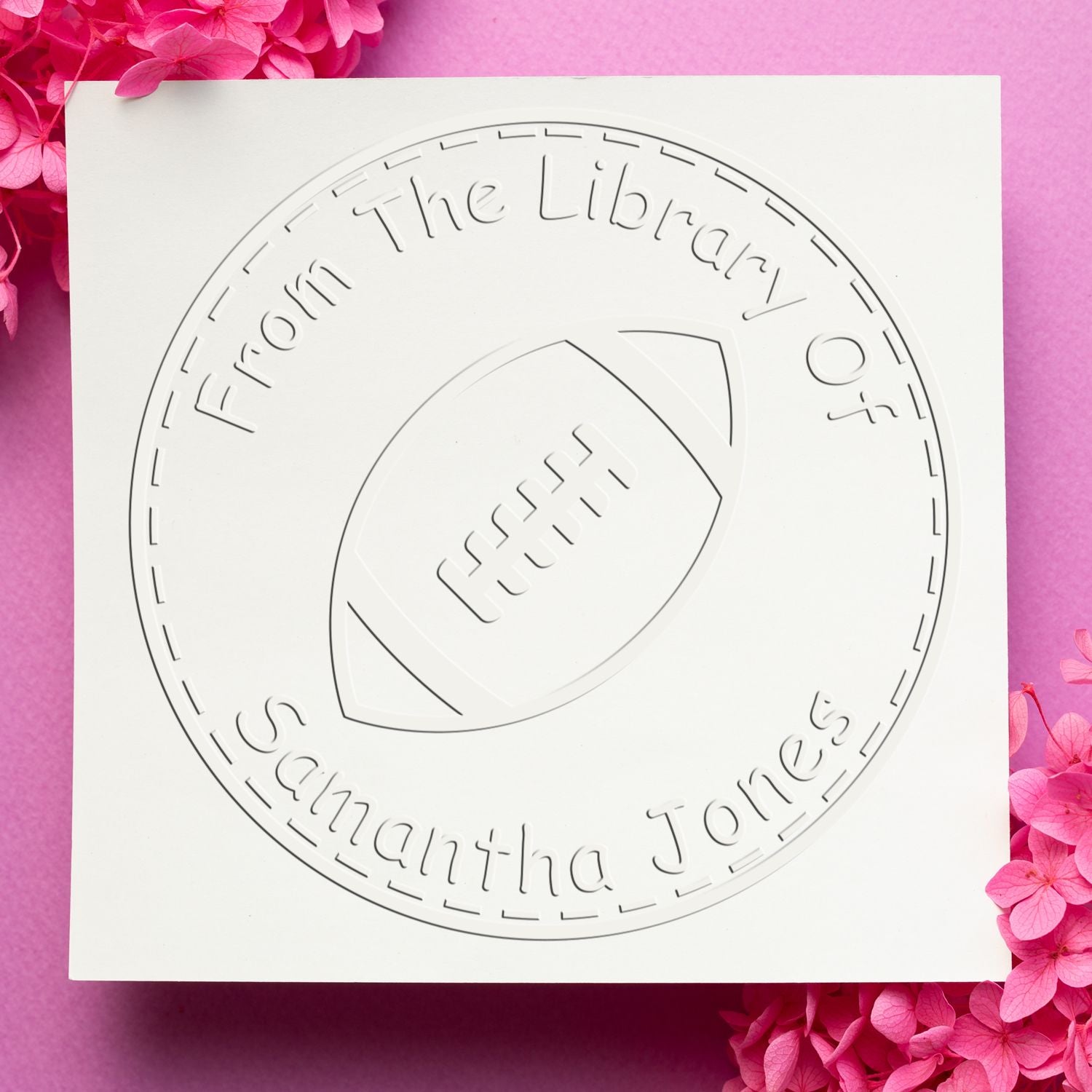 White circular football stamp and seal product with embossed text, on a pink background with bright pink flowers.
