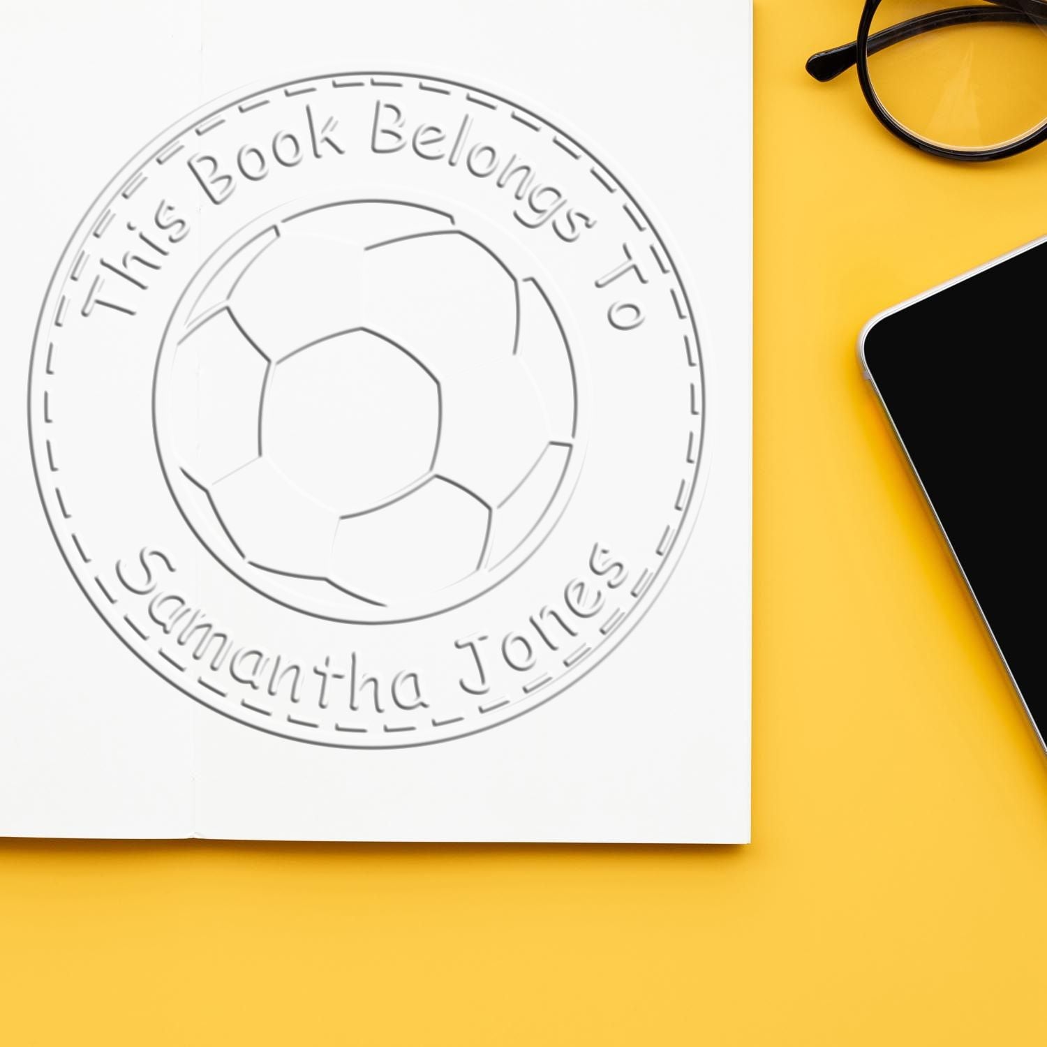 Custom book ownership stamp seal with soccer ball, embossed white circular design on a yellow background beside glasses and phone.