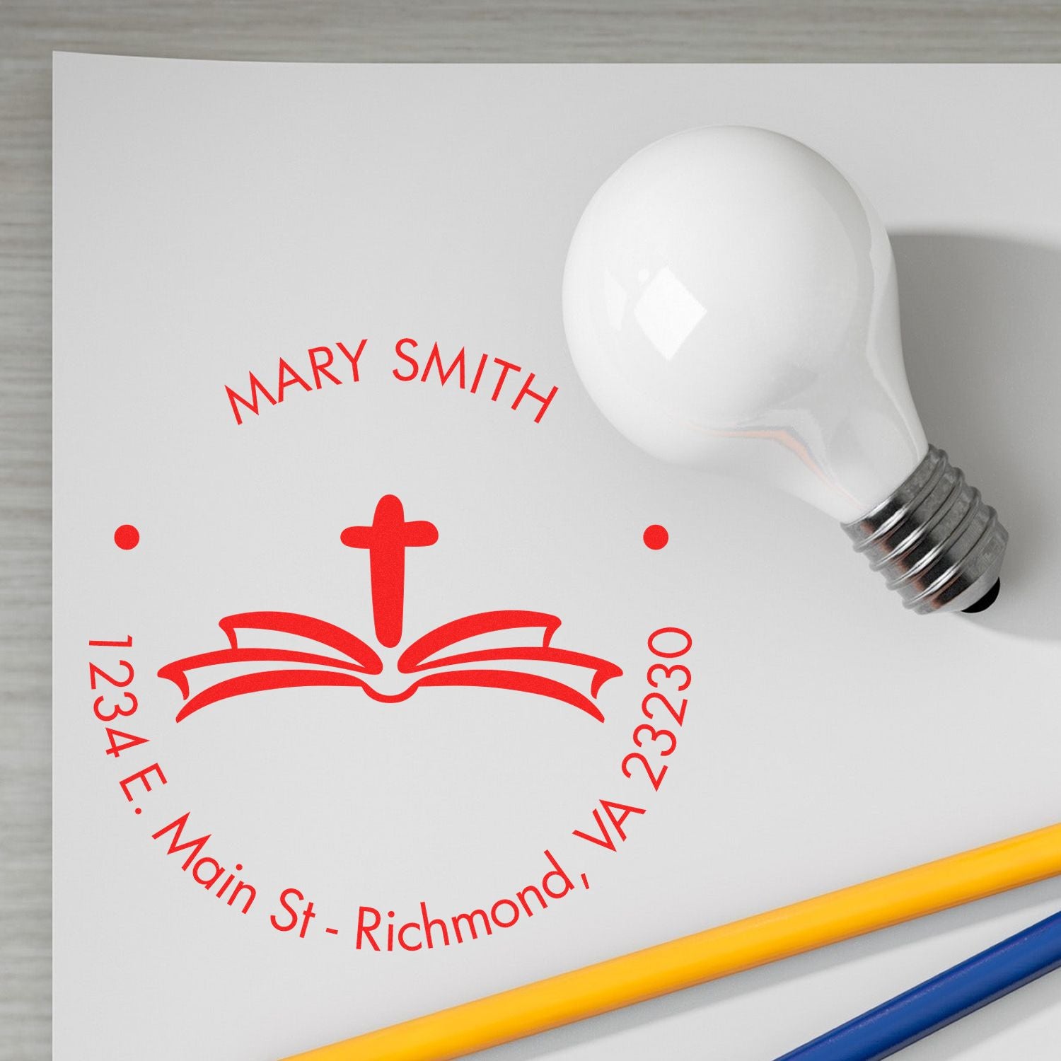 Red custom stamp and seal with Mary Smith, cross and book logo, on white paper beside a white light bulb.