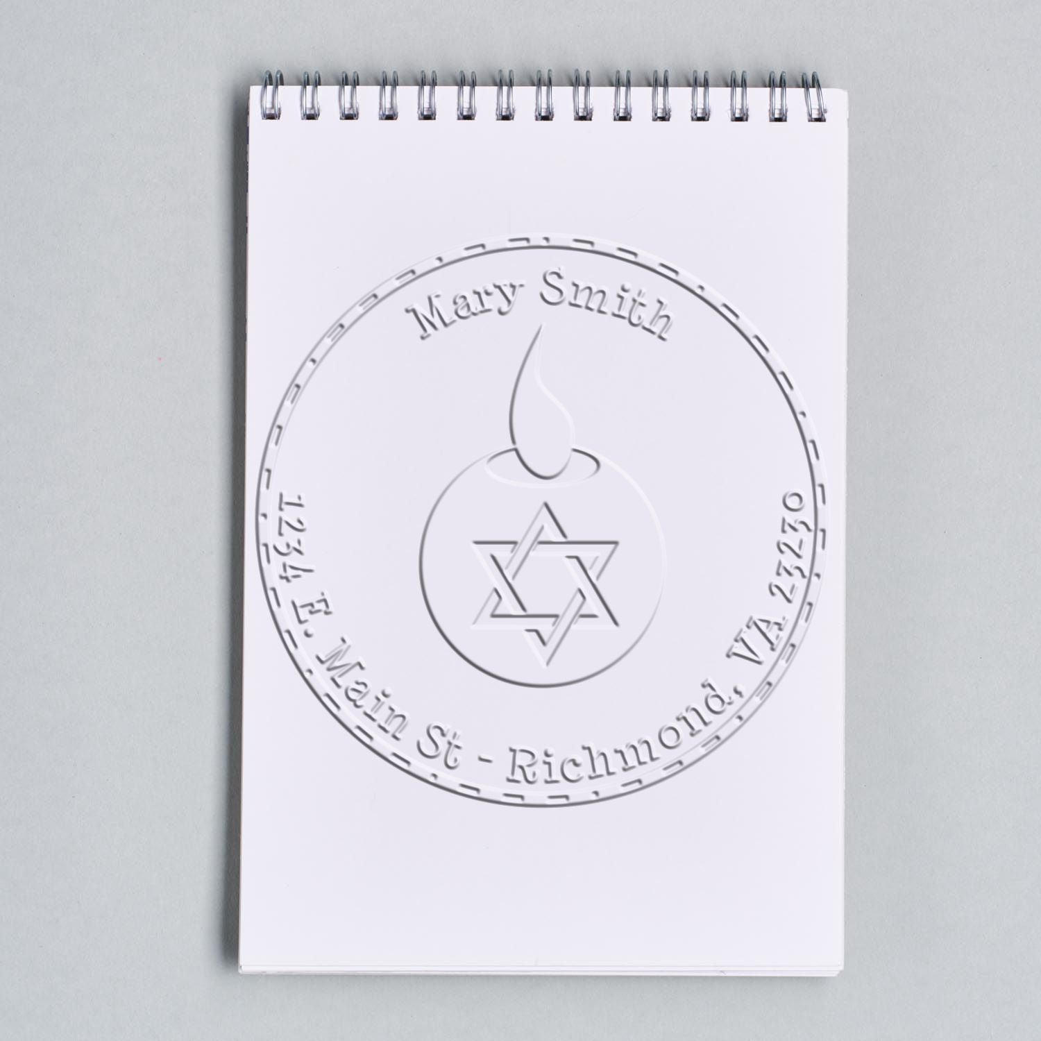 White spiral notepad featuring an embossed gray custom stamp seal with circular border, candle icon, and Star of David.
