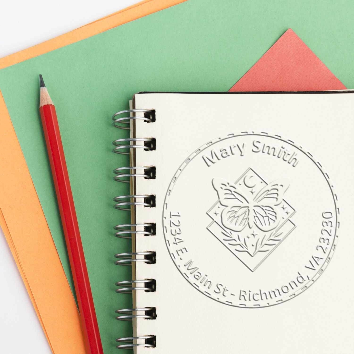 Custom butterfly embossing seal on a spiral notebook, with a red pencil and green-orange paper.